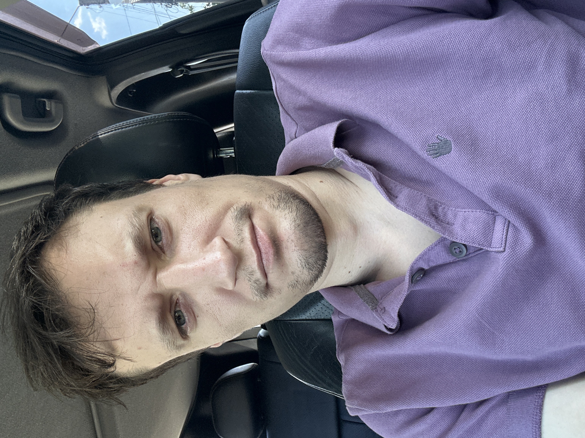 A male individual is captured in a close-up, medium-shot selfie taken inside a vehicle. He is positioned in the driver's seat, facing the camera directly. The individual has medium-length brown hair, styled somewhat messily. His facial expression appears somewhat strained or concerned, with his eyebrows furrowed. He has light-colored eyes and stubble on his chin and upper lip. He is wearing a light purple polo shirt. The interior of the car is visible, with the headrest of the passenger seat in the background, as well as parts of the car's ceiling and door frame. The outdoor environment is partially visible through the car window, showing a bright sky with some clouds and indistinct structures. The image is well-lit, suggesting it was taken during the daytime. The specific location of Makiv, Ukraine, is not discernible from the visual elements within the image itself.