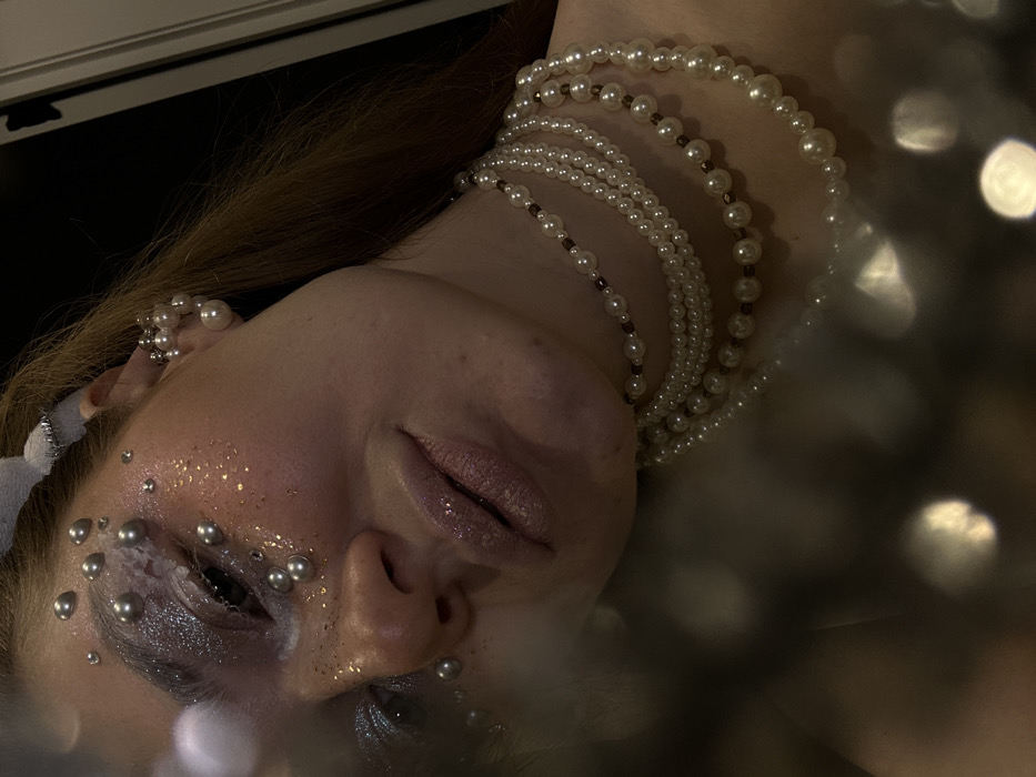 A partial portrait depicts a female subject in Kazan, Russia, gazing slightly upwards with her head tilted. Her left eye is visible, accentuated with silver glitter eyeshadow, small white crystalline embellishments, and numerous spherical silver-colored beads adhered to the skin around the eye, cheekbone, and upper temple. Gold glitter is scattered on the left cheek. Her lips are slightly parted, coated in a glossy, light pink substance. Reddish-brown hair is visible on the right side of her head, along with a white, possibly fabric, hair accessory. The subject wears a cluster earring of white spherical beads on her visible left ear. Multiple layered necklaces, primarily composed of white imitation pearls, are draped around her neck; some strands integrate smaller, dark brown cylindrical beads between the pearls. The background is dark and out of focus, featuring a vertical light-colored edge on the far right. The lower left foreground contains large, blurred, circular light reflections.