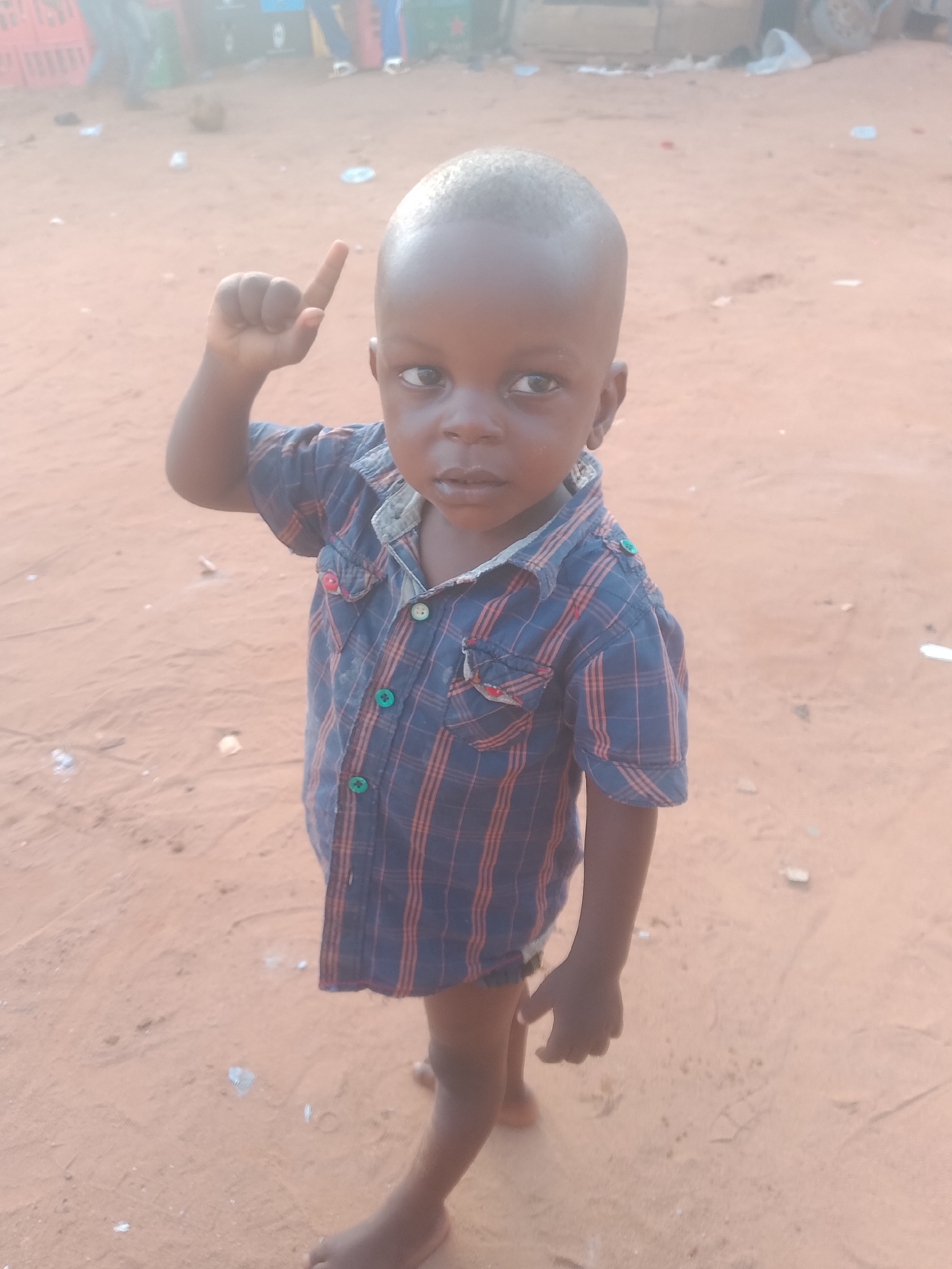A young child stands barefoot on a sandy surface in Nkpor, Nigeria. The child, possibly a toddler, has dark skin and is wearing a blue and orange plaid shirt that is only partially buttoned. The child raises its right index finger. In the background, indistinct shapes suggest the presence of crates and other objects.