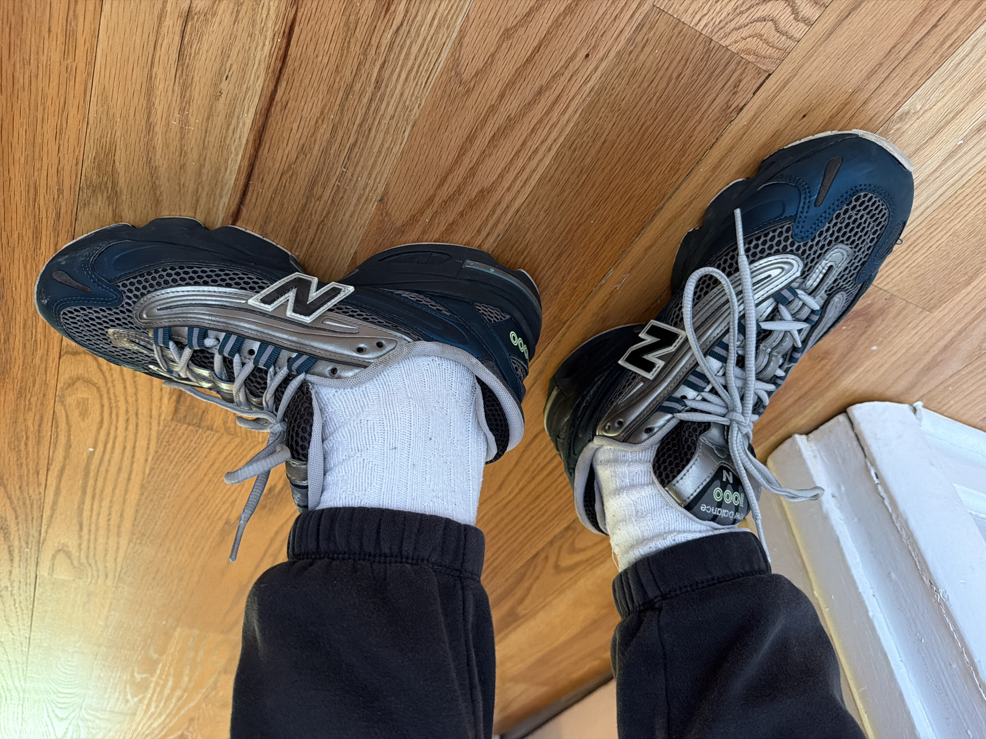The image shows a person wearing New Balance 1000 sneakers, grey socks, and dark-colored sweatpants, standing on a wooden floor. The sneakers are primarily navy blue and silver with grey laces and have the New Balance "N" logo on the sides. The photo is taken from an overhead perspective, looking down at the person's feet. The location appears to be indoors, potentially a home, and the wooden floor is light brown. While the location is in the United States of America, it cannot be determined that the location is specifically in the city of Morton Grove.