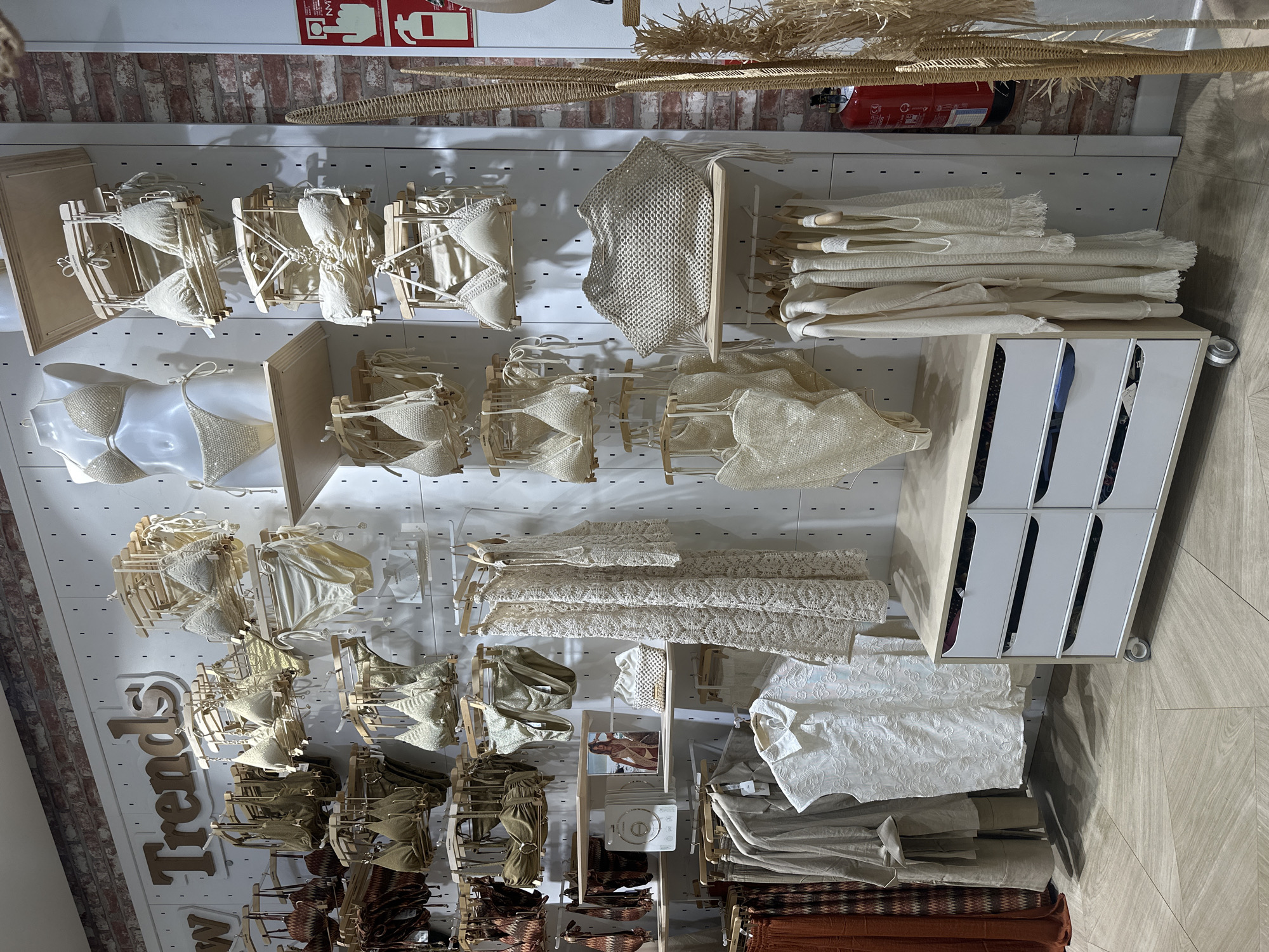 A display of clothing hangs on a wall, with various items, including swimwear, dresses, shirts, and trousers. The clothes are generally in neutral tones. The wall is white with a grid pattern, and shelves support the garments. To the right, a white cabinet with drawers is visible. There is a fire extinguisher on the upper right, mounted on a brick wall. Text reads "New Trends" is visible on the lower left.