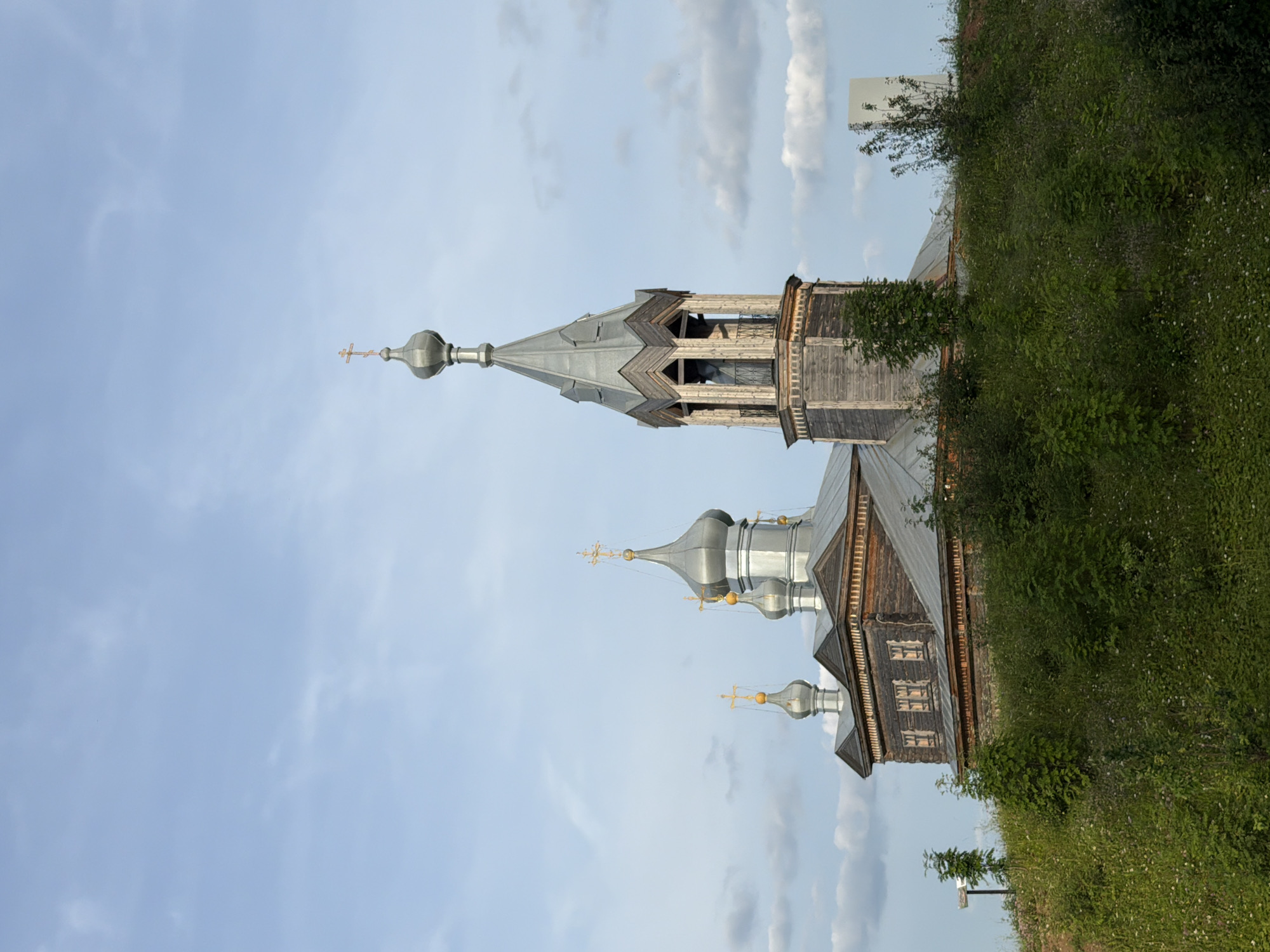 A wooden Orthodox church with multiple domes and a bell tower stands on a grassy hillside under a cloudy blue sky. The church features silver domes topped with gold crosses. The main structure is made of dark wood, with smaller windows visible on the side. The bell tower is tall and octagonal, also constructed from wood, with open sections revealing its interior. The foreground is filled with dense green vegetation, including trees and wildflowers. In the distance, a plain white rectangular structure is visible behind some trees. The scene appears to be in Cherdyn, Russia, given its distinct architectural style commonly found in the region. There are no people or animals visible in the image.