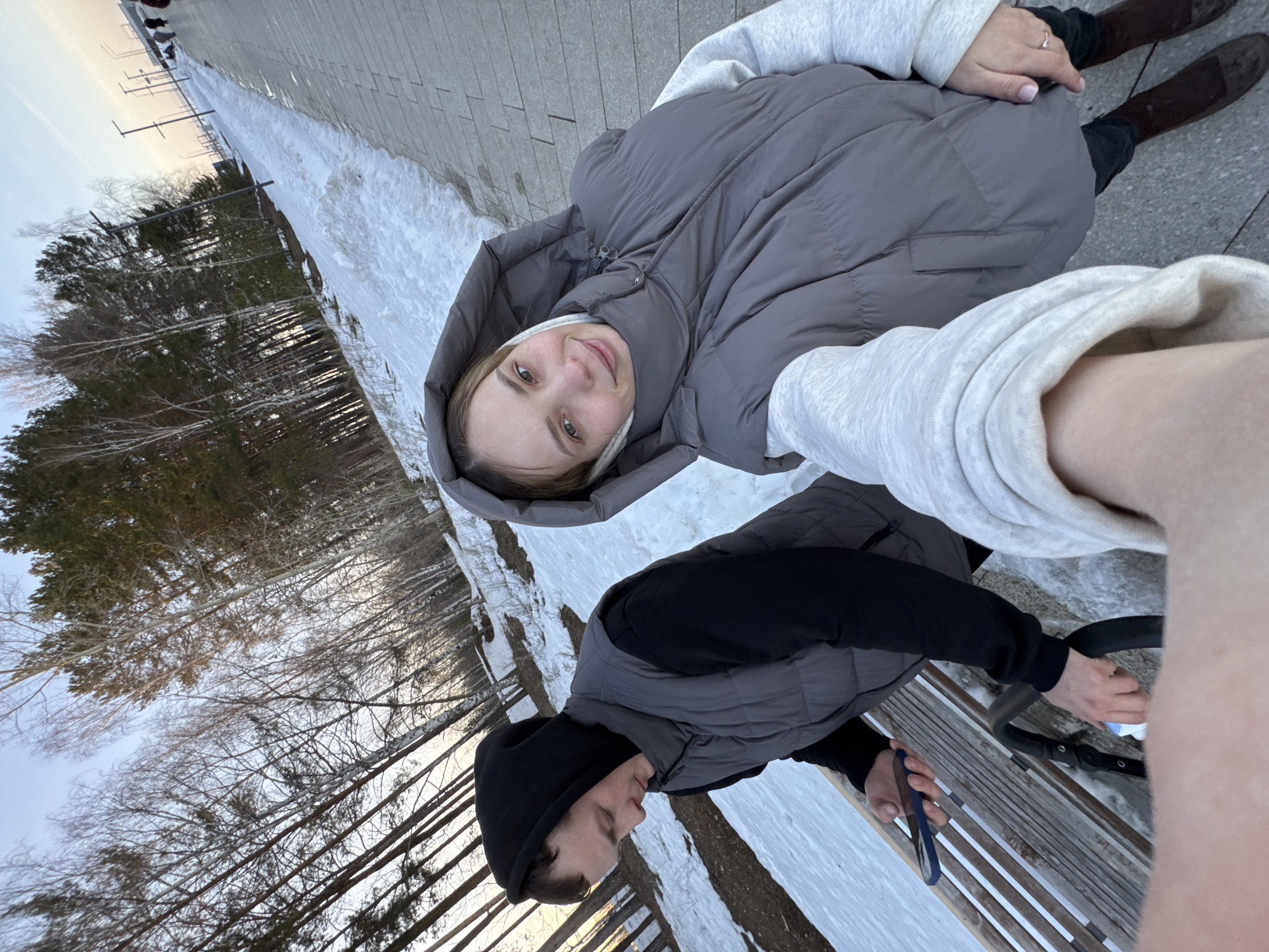 Two people are positioned in the foreground of a photograph taken in Chaikovsky, Russia. A woman wearing a gray hooded puffer vest over a light-colored sweatshirt is looking at the camera. Seated next to her is a man wearing a black beanie and a gray puffer vest over a black long-sleeved shirt. He is holding a blue phone in his hand. They are sitting on a bench. The ground adjacent to the bench has patches of snow. In the background are trees without leaves. A pathway is also visible.