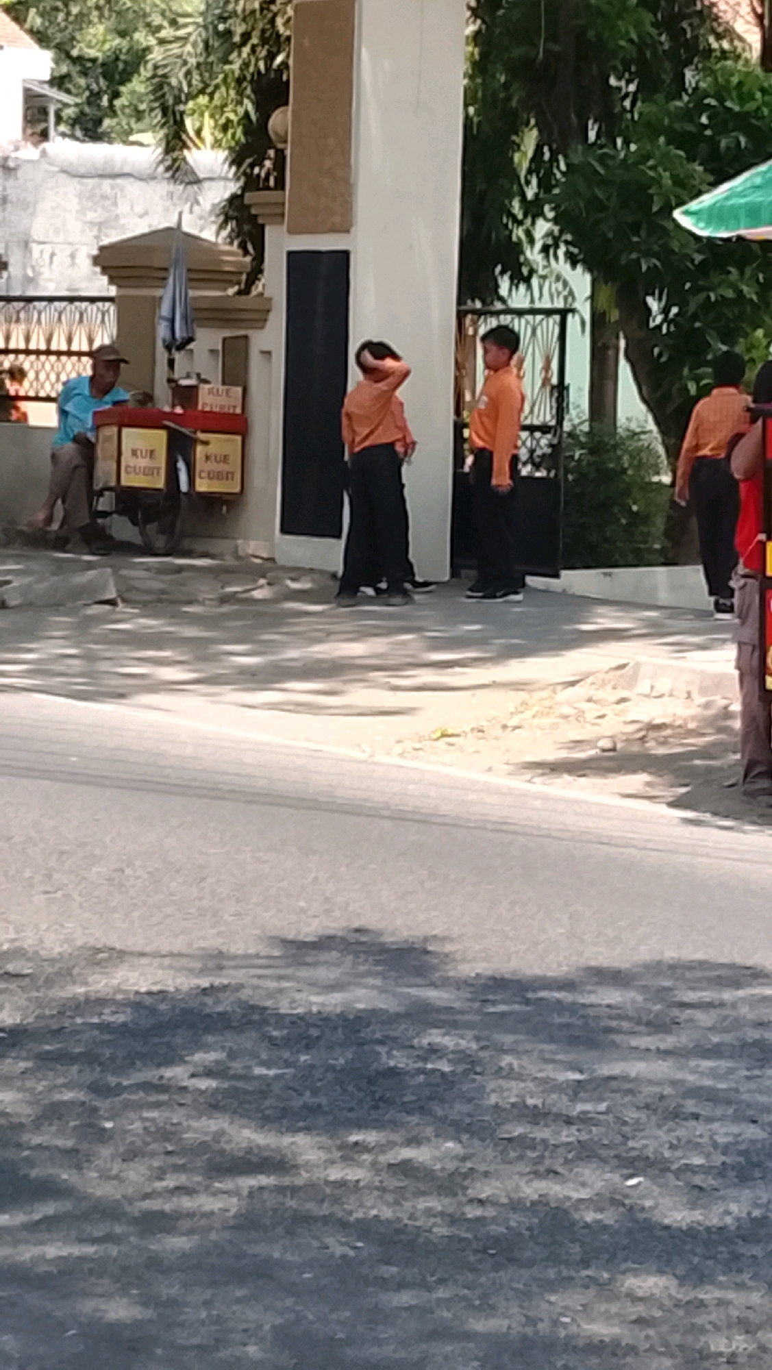 The image captures a street scene in Slawi, Indonesia, likely during the daytime given the bright sunlight casting shadows. In the foreground, a paved road with distinct tire tracks is visible, with dappled sunlight falling across it. To the left of the frame, a man wearing a blue shirt and a brown hat is seated near a food cart. The cart has labels that read "KUE CUBIT" and appears to be a mobile vendor. An umbrella is resting on the cart. Behind the cart, a white pillar forms part of a building's facade, with a black panel.
To the right of the pillar, two young individuals in orange shirts and dark pants are standing. One is facing away from the camera, with his hand raised to the back of his head. The other is facing to the side, also in the orange shirt and dark pants. Further to the right, another person in an orange shirt and dark pants is visible, partially obscured, and appears to be walking away from the scene. A glimpse of a green and white umbrella is visible in the upper right corner, suggesting a market or food stall area. Trees and foliage are present in the background, indicating an outdoor setting within the city. The overall impression is a candid snapshot of everyday life on a street in Slawi.