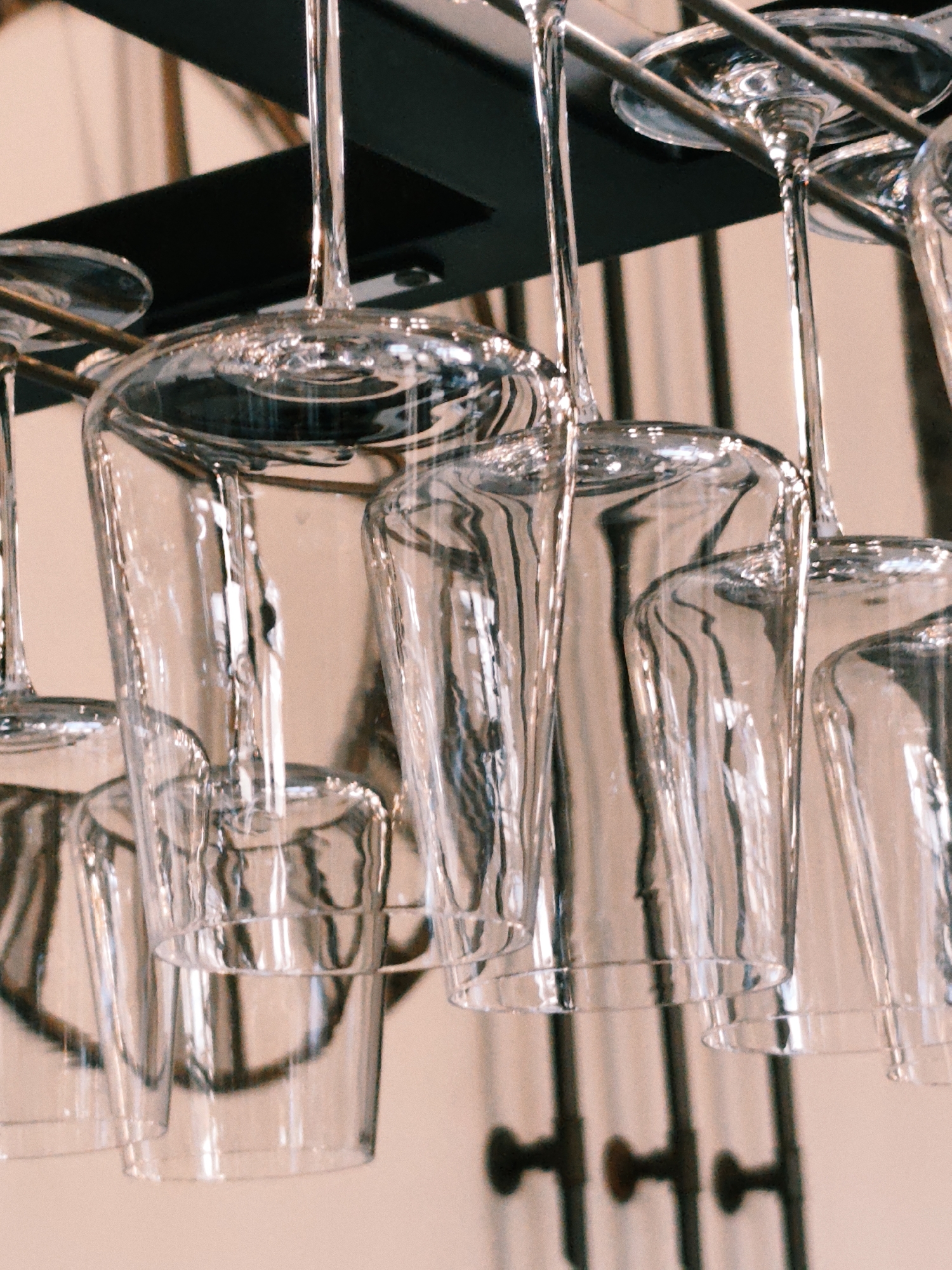 The image shows a close-up of several clear wine glasses hanging upside down from a black rack. The glasses are made of glass and have long stems. The background is a blurred peach-colored wall with vertical lines, possibly suggesting vertical blinds or a slatted structure. The lighting creates reflections and highlights on the glass surfaces. The scene appears to be indoors, possibly in a restaurant or bar setting in Estosadok, Russia, given the implied location. There are no people visible in the image, and the focus is solely on the arrangement of the glassware.