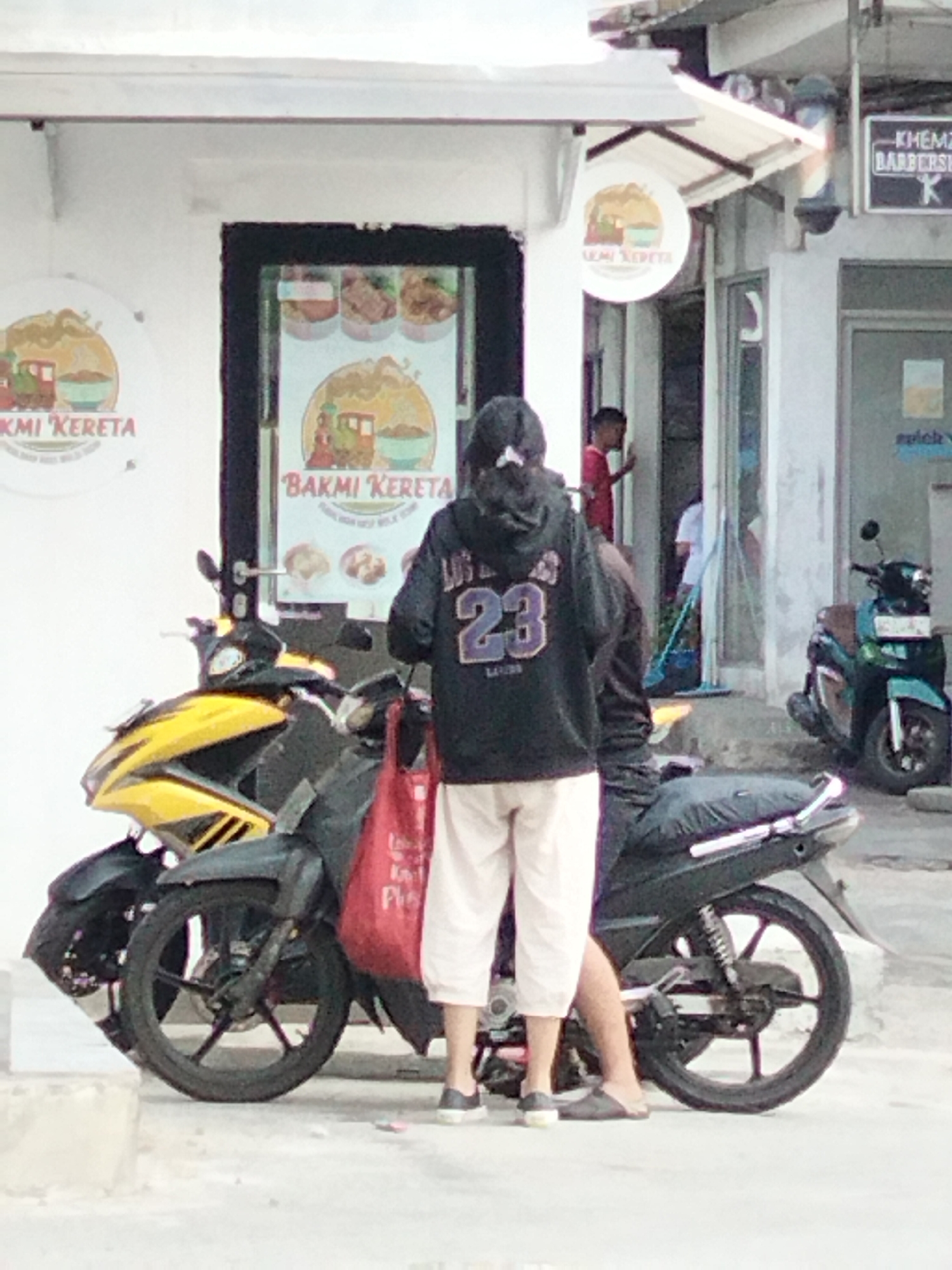 This is a medium shot image taken outdoors, depicting a street scene in South Tangerang, Indonesia, likely near a commercial establishment. In the foreground, two motorcycles are parked. The motorcycle on the left is yellow and black, while the one on the right is predominantly black. A person wearing a black hoodie with the number "23" on the back and light-colored pants is standing next to the black motorcycle, holding a red bag. Behind the person, a young boy in a red shirt is visible near the entrance of a building. The building appears to be a shop or restaurant, with signage that includes "BAKMI KERETA." Another teal-colored motorcycle is parked to the right of the entrance, and a barber pole is visible further down the street, indicating the presence of a barbershop. The ground is paved and appears to be a sidewalk or street. The overall impression is a casual urban scene with elements of commerce and transportation.