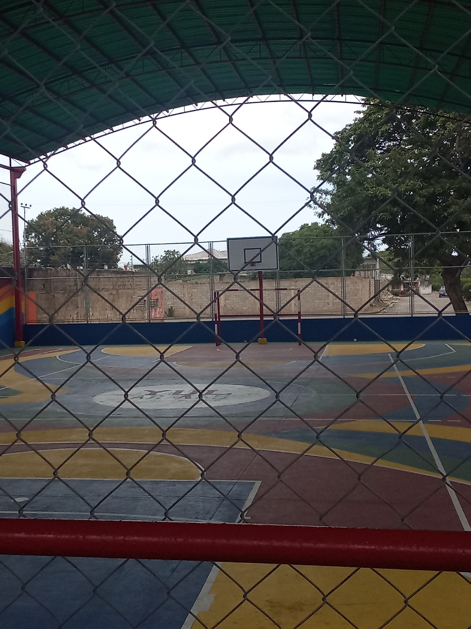 A fenced-in basketball court, with an overhead structure, is seen through a chain-link fence. The court has multiple painted colors. A backboard and goal stand prominently. Trees are seen outside the fenced area. The location is most likely in Juan Griego, Venezuela.
