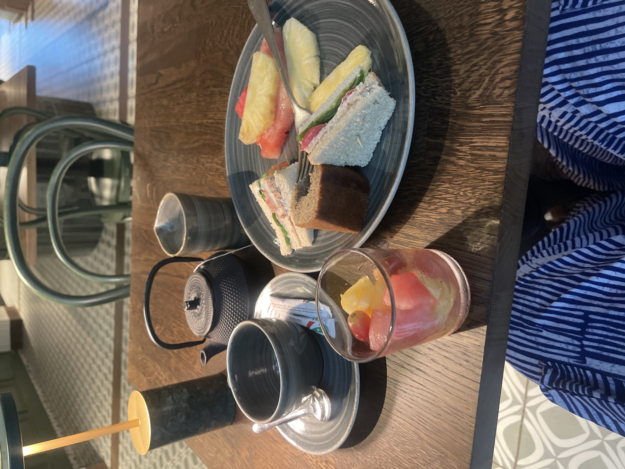 The image shows a table setting, likely inside a cafe or restaurant. The table is made of wood. There is a plate with finger sandwiches, pineapple slices, and watermelon. A fork is stuck into one of the sandwiches. A teapot, cup, and small glass filled with fruit are also on the table. A patterned shirt is visible in the background. The image was not taken in Abuja, Nigeria.