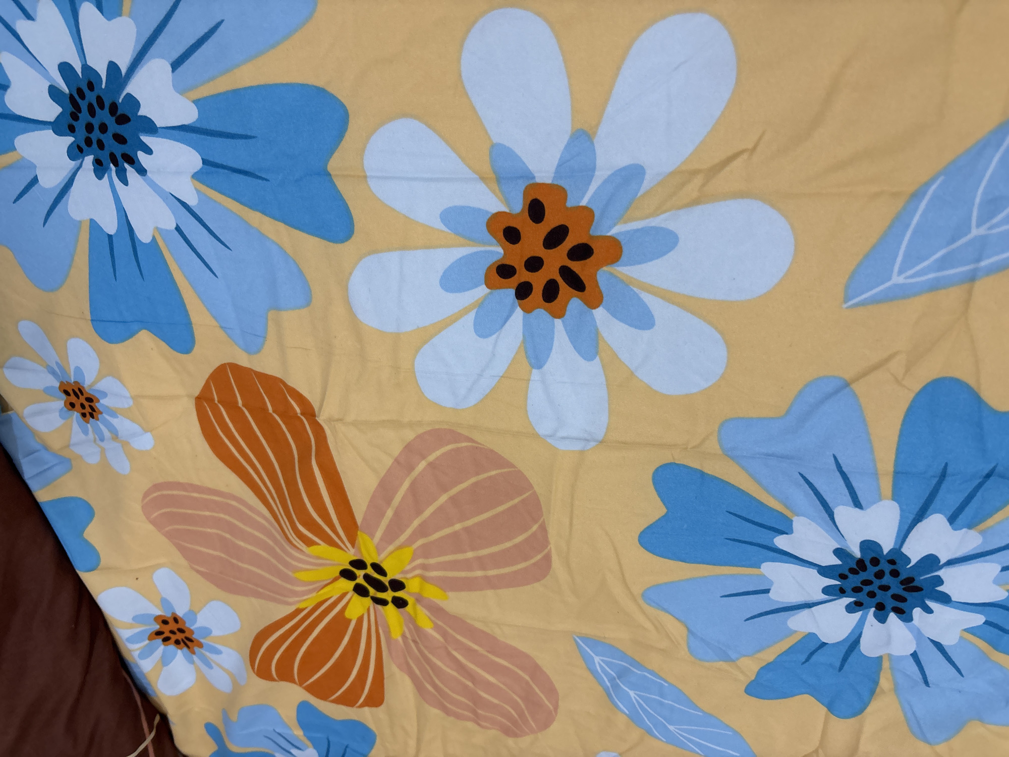 A close-up, slightly angled shot shows a yellow bedsheet with a floral pattern. The flowers are depicted in a stylized, graphic manner, with bold outlines and flat colors. There are large blue and white flowers with orange centers, and a large orange and yellow flower with a black center. Blue leaves with white veins are scattered across the bedsheet. The fabric is slightly creased, suggesting it is either a duvet cover or a fitted sheet. The top left of the image shows a corner of a darker brown, possibly upholstered surface, like a headboard, angled over the bedsheet. The scene does not contain any identifiable elements that would suggest a specific location in Wanajaya, Indonesia, or any other city.
