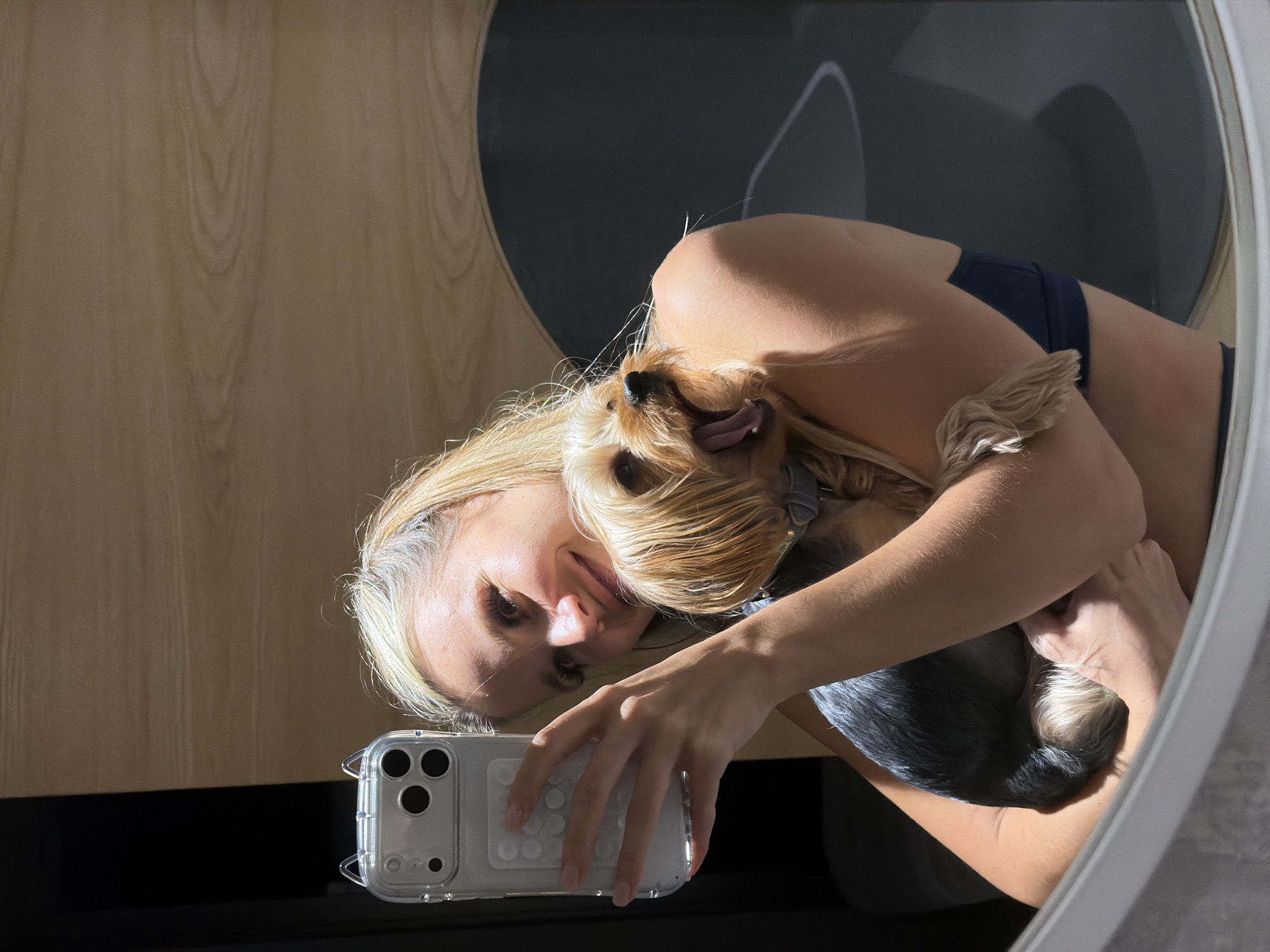 A blonde woman takes a mirror selfie while holding a Yorkshire Terrier. The woman is wearing a dark blue bra and her hair is pulled back. The dog is held in her arms, with its mouth open as if yawning or barking. The background appears to be indoors with wooden paneling and a large, dark circular object. The setting is not specified as being in Thep Krasatti, Thailand.