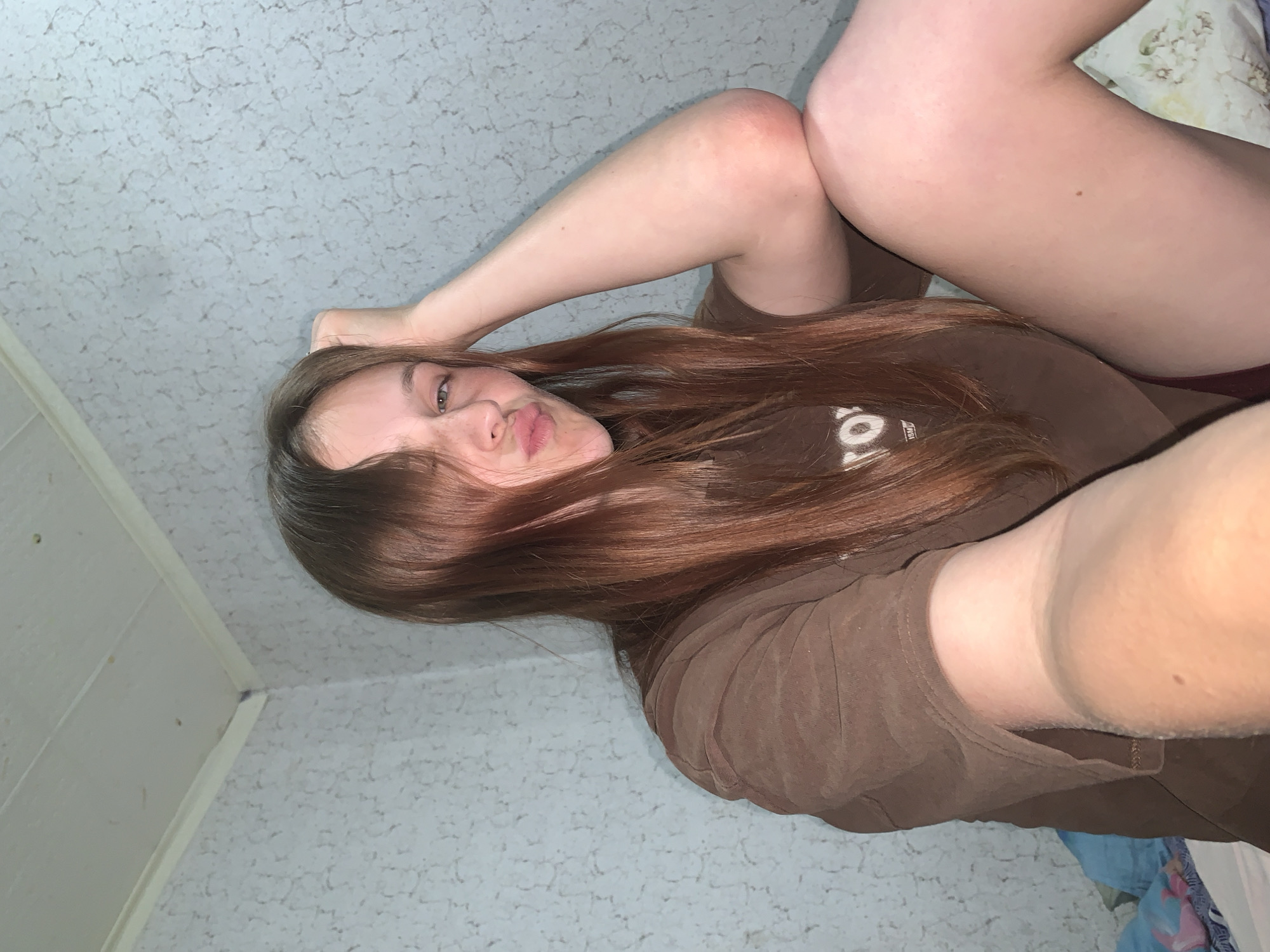 A young woman is captured in a selfie from a high angle, looking down as she lies or reclines. She has long, straight, reddish-brown hair that cascades over her shoulders and partially covers her forehead. Her eyes are light-colored, and she poses with puckered lips in a playful expression. She wears a plain brown t-shirt with partially visible white text on the chest, appearing to include letters such as 'Y' and 'O'. Her bare legs are bent, with one knee showing a distinct reddish mark or scrape. The background is predominantly a light-colored, heavily textured ceiling, characteristic of a popcorn or stucco finish. In the upper right corner, a small section of a patterned pillow or blanket is visible. To the left, a corner where the ceiling meets a light-colored wall, possibly featuring vertical paneling, can be seen. The indoor lighting suggests a home environment. The image conveys a casual and spontaneous moment, likely taken in a residence in Фоки, Russia.