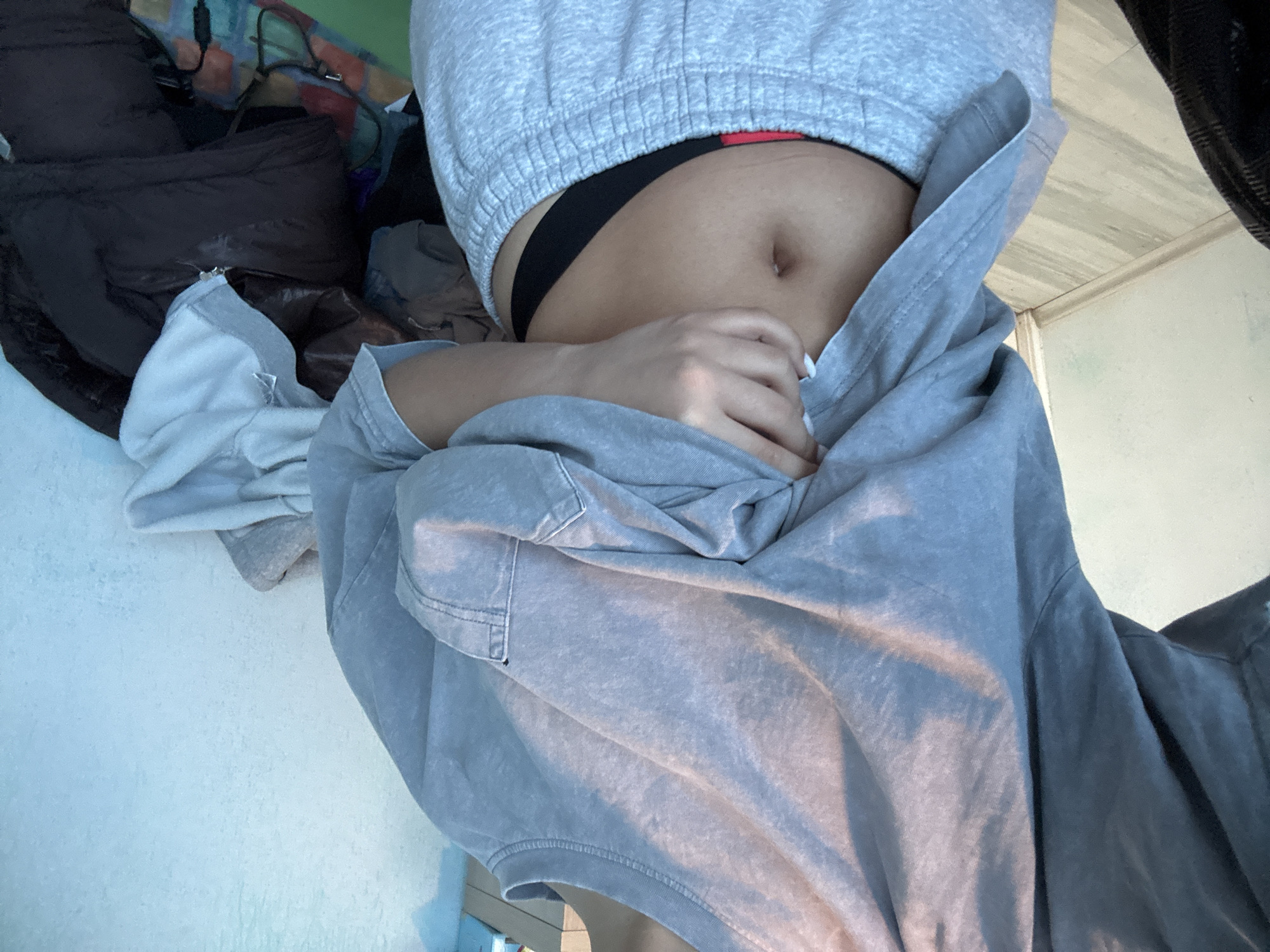 A close-up, partially obscured, seemingly low-angle shot depicts a person's torso and hand against a cluttered background. The person is wearing a light gray hooded sweatshirt that is pulled down to expose their abdomen, revealing a black thong. Their hand covers part of their stomach. The surrounding environment consists of various items, including a dark-colored bag and assorted clothing items, suggesting a cluttered space. The scene is lit unevenly, with lighter areas contrasting with shadowed sections.
