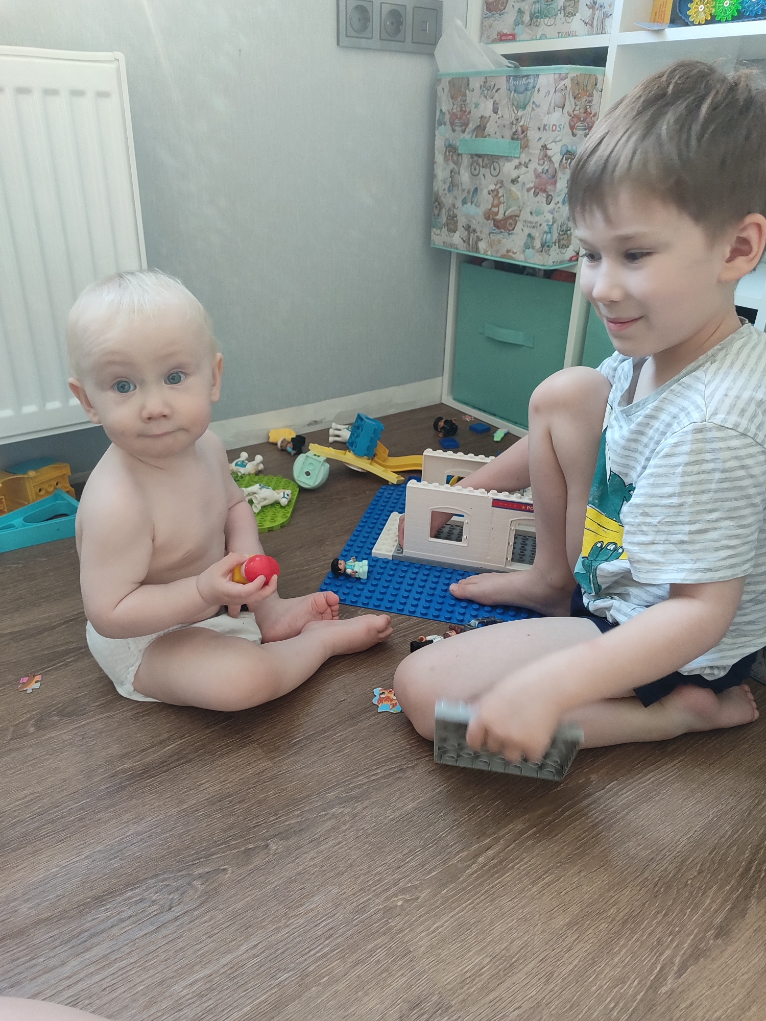 Two young children are sitting on a wood-look floor in a room. A fair-skinned baby, wearing a diaper, is seated on the left holding a red and yellow toy. To the right, a fair-skinned older child with short brown hair is seated cross-legged and is playing with LEGO bricks. The older child is wearing a striped t-shirt with a graphic design. Various LEGO pieces and small toys are scattered around them. A white radiator is visible against a light gray wall on the left. A white shelving unit with decorative storage bins is in the background. A light switch is visible on the wall. The location is likely indoors in Izhevsk, Russia, given the information provided.
