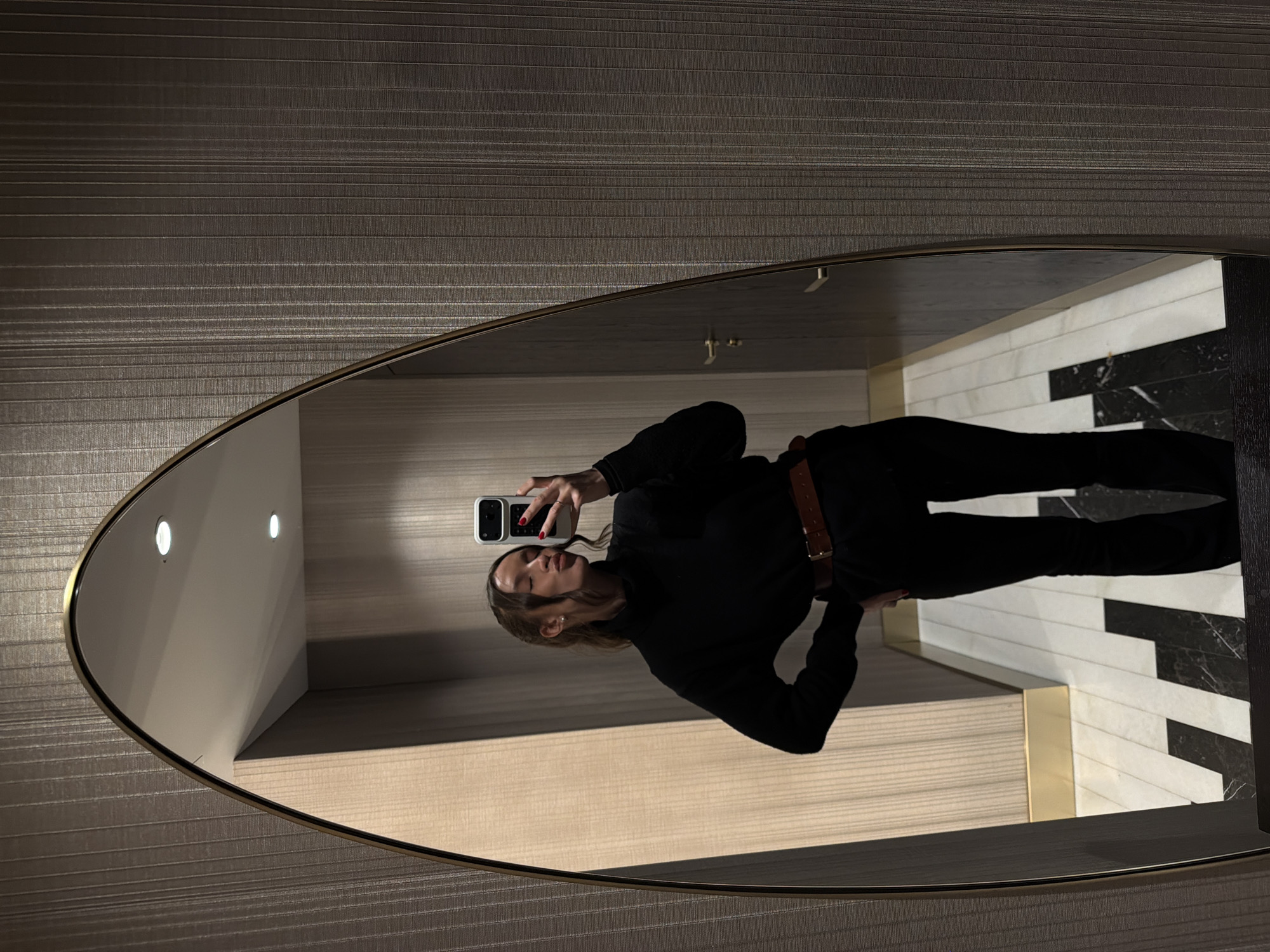 The image depicts a mirror selfie taken indoors within a contemporary setting in Budva, Montenegro. A single adult female subject is reflected in a large, irregularly shaped mirror featuring a thin gold-toned frame. She is dressed in a black turtleneck top, black trousers, and a brown belt. Her left hand is placed on her hip, while her right hand holds a white-cased smartphone, elevated to capture the reflection. The subject's head is tilted, her eyes are closed, and her lips are pursed with the tongue slightly extended. Red nail polish is visible on her left hand.
The background reflected in the mirror reveals an interior hallway or lobby characterized by textured wall coverings. A dark gray or brown wallpaper with vertical striations is visible on the primary wall. Another wall section, appearing lighter in color, also features a vertical linear texture. The floor exhibits a distinct geometric pattern of alternating black and white marble or tile segments. Two circular recessed lights illuminate the ceiling. A dark-colored door with a gold-toned handle is present in the reflected background, alongside a gold-colored baseboard at the wall-floor junction.