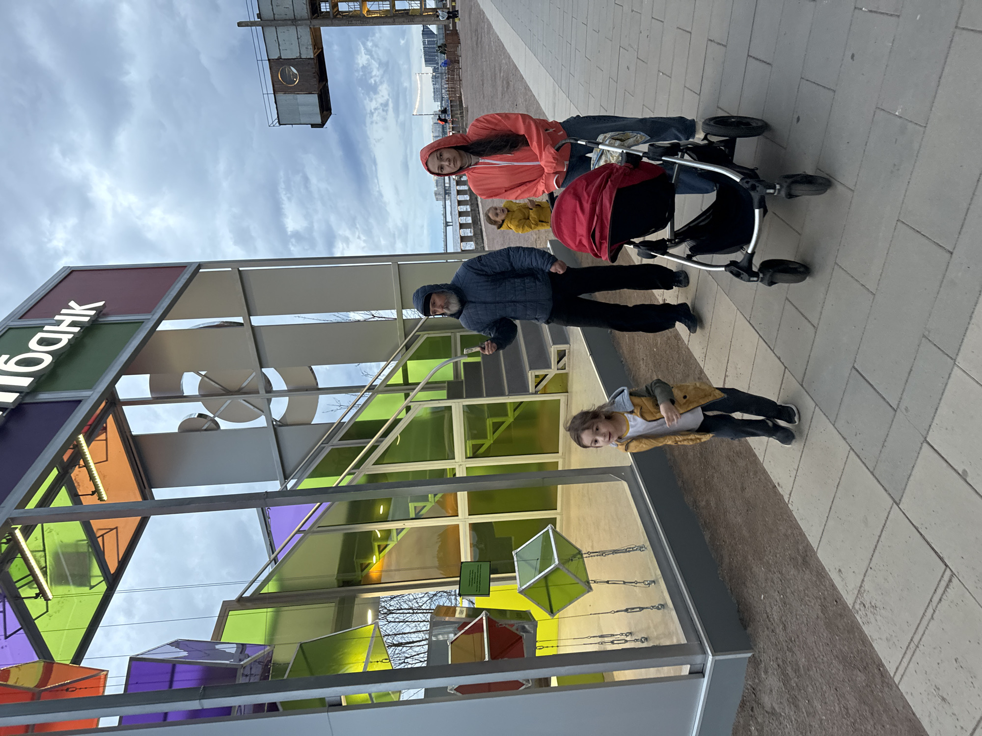 A man and a woman with a stroller are walking on a paved pathway in Saint Petersburg, Russia. The man, wearing a blue puffer jacket and black pants, holds a smartphone in his right hand. The woman, in a red hooded jacket and blue jeans, pushes the stroller. A young child in a yellow jacket stands behind them. To the left of the pathway is a modern structure with colored glass panels and stairs, which appears to be an art installation or part of a building. The background shows a body of water, a bridge, and buildings, characteristic of Saint Petersburg. The sky is overcast with clouds.