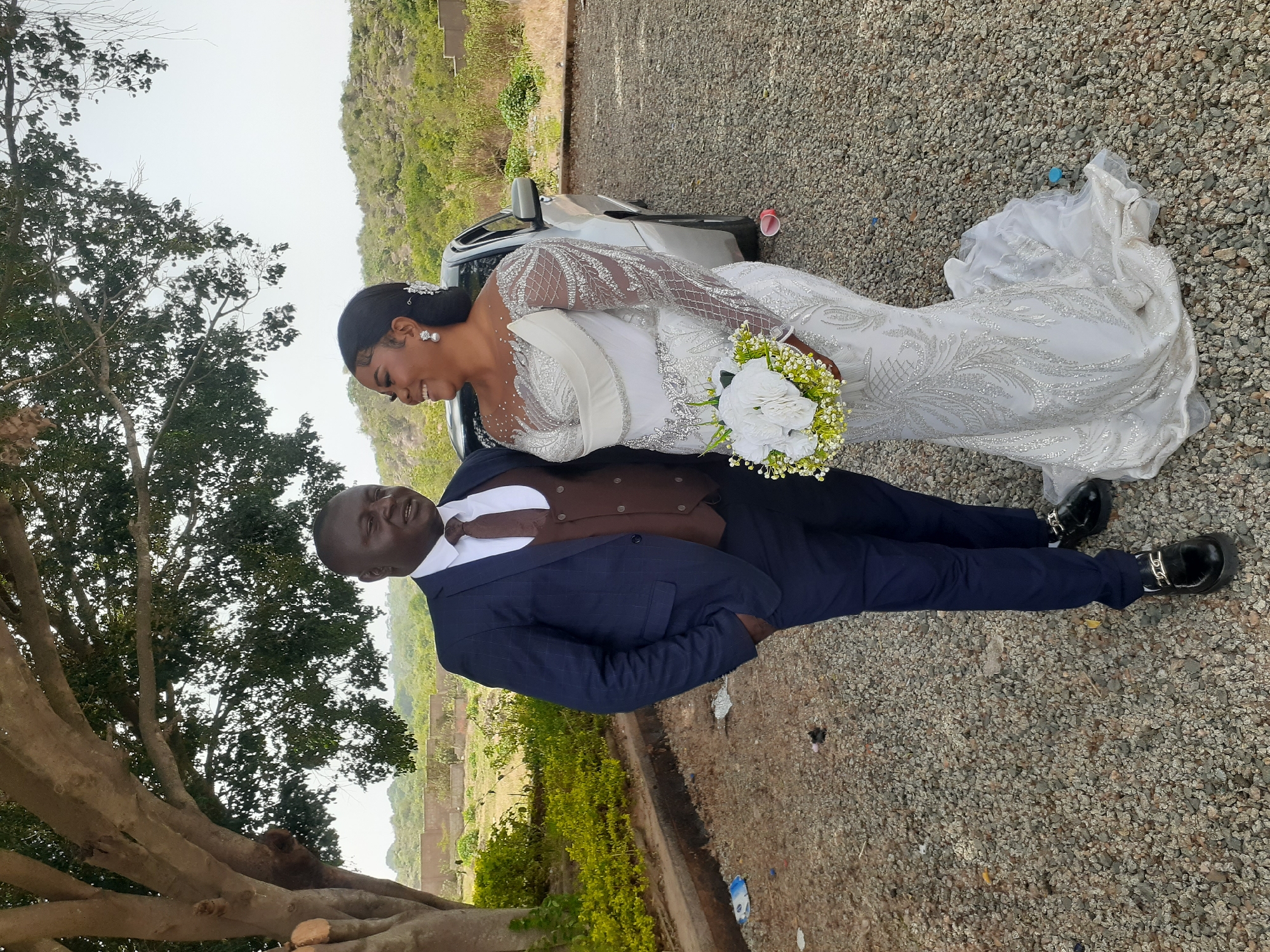The image captures a wedding scene in Jos, Nigeria. A bride, wearing a long-sleeved, embellished white gown, stands beside a groom. The groom is in a navy blue suit with a brown vest and tie. They are standing on a gravel ground near a car, against a backdrop of a landscape with trees and hills. A large tree is visible on the left side of the image.