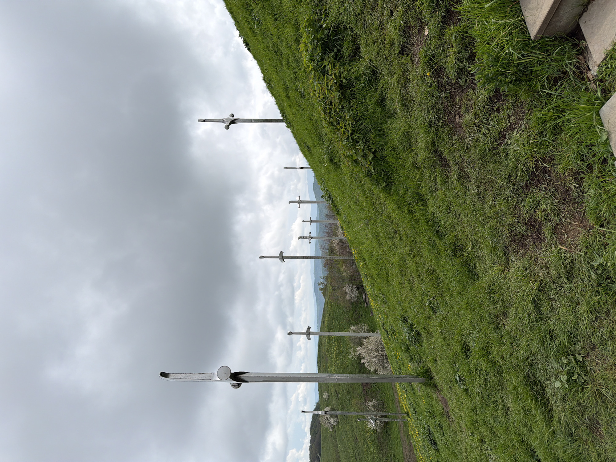 The image shows a line of large, sword-shaped monuments extending across a grassy hillside towards the sky. The monuments are made of a silver or gray material. The hillside is covered in green grass and wildflowers. There is a cloudy sky above. The location is Zemo Nichbisi, Georgia.
