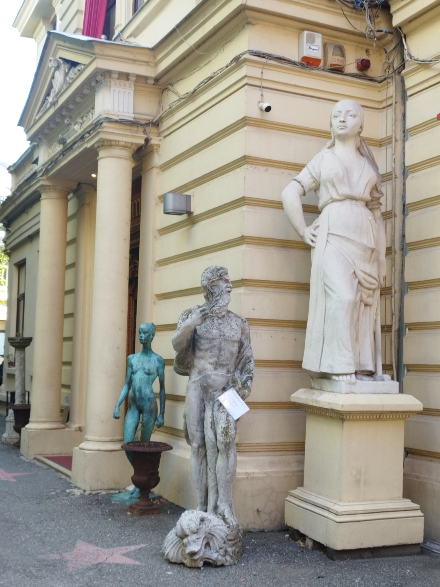 This image depicts a scene outside a building, likely a museum or historical site, in Bucharest, Romania. In the foreground, several statues are displayed on the pavement and on pedestals. A large, weathered stone statue of a muscular man with a beard stands prominently in the center. To its left, a smaller, teal-colored bronze statue of a nude woman stands on a low pedestal. Further to the left, a third statue, also of a nude woman and made of white stone, is positioned on a taller, ornate pedestal. The building itself is classical in design, with large columns and a cream-colored facade adorned with decorative elements and horizontal banding. Two security cameras are visible on the building's exterior, along with what appear to be alarm system components. The ground is paved with asphalt, and the scene appears to be outdoors during daylight.