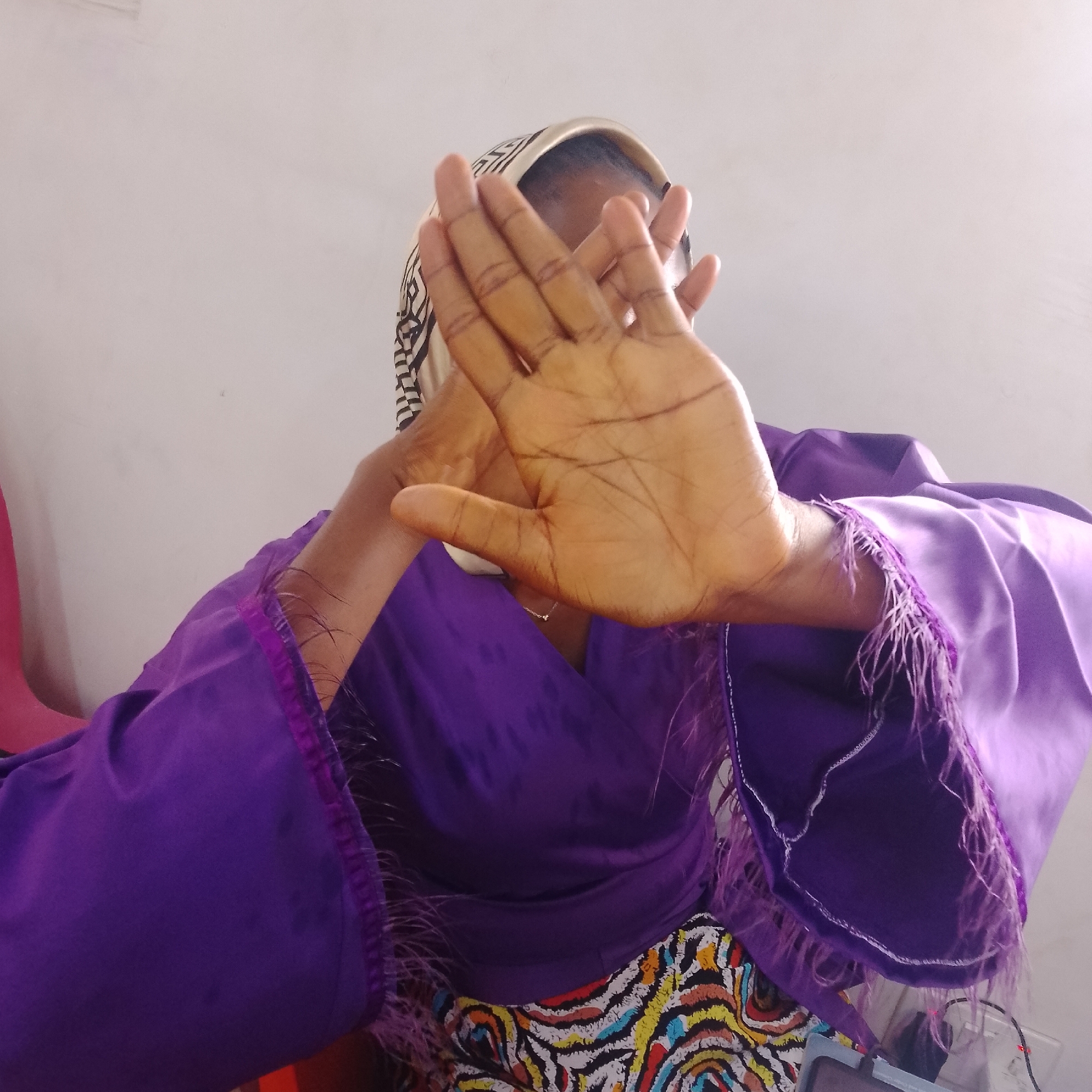 In a room with plain white walls, a person wearing a purple headscarf and a vibrant purple robe with fringed edges holds their hands up to block the camera. The robe has a patterned trim with black, white, yellow, blue, and orange colors. The person's hands are large and visible, with lines on the palms. The background is out of focus, suggesting the image was taken indoors. The image does not provide specific information about the location within Iranla, Nigeria, beyond being an indoor setting.