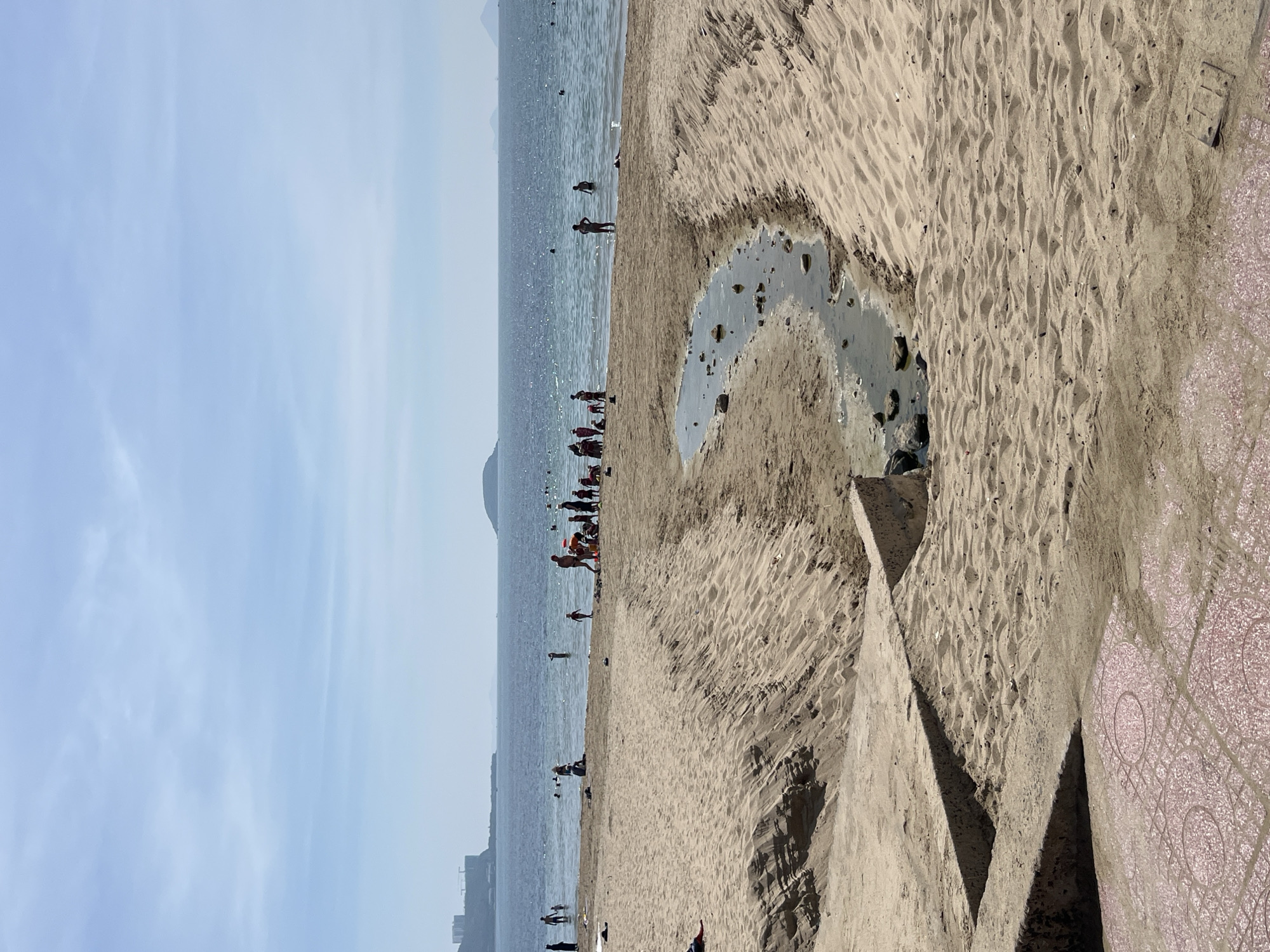 The image depicts a crowded beach scene in Nha Trang, Vietnam, under a clear sky with scattered clouds. In the foreground, a sandy embankment slopes down towards the sea, with a small, somewhat murky stream flowing across it. Some concrete structures are visible along the embankment. The beach is populated with numerous people, many of whom are wading in the shallow water or gathered in groups on the sand. Some individuals are sunbathing. In the background, the ocean stretches towards a distant coastline with several small islands and a hazy cityscape. The overall impression is of a lively, popular beach on a sunny day.