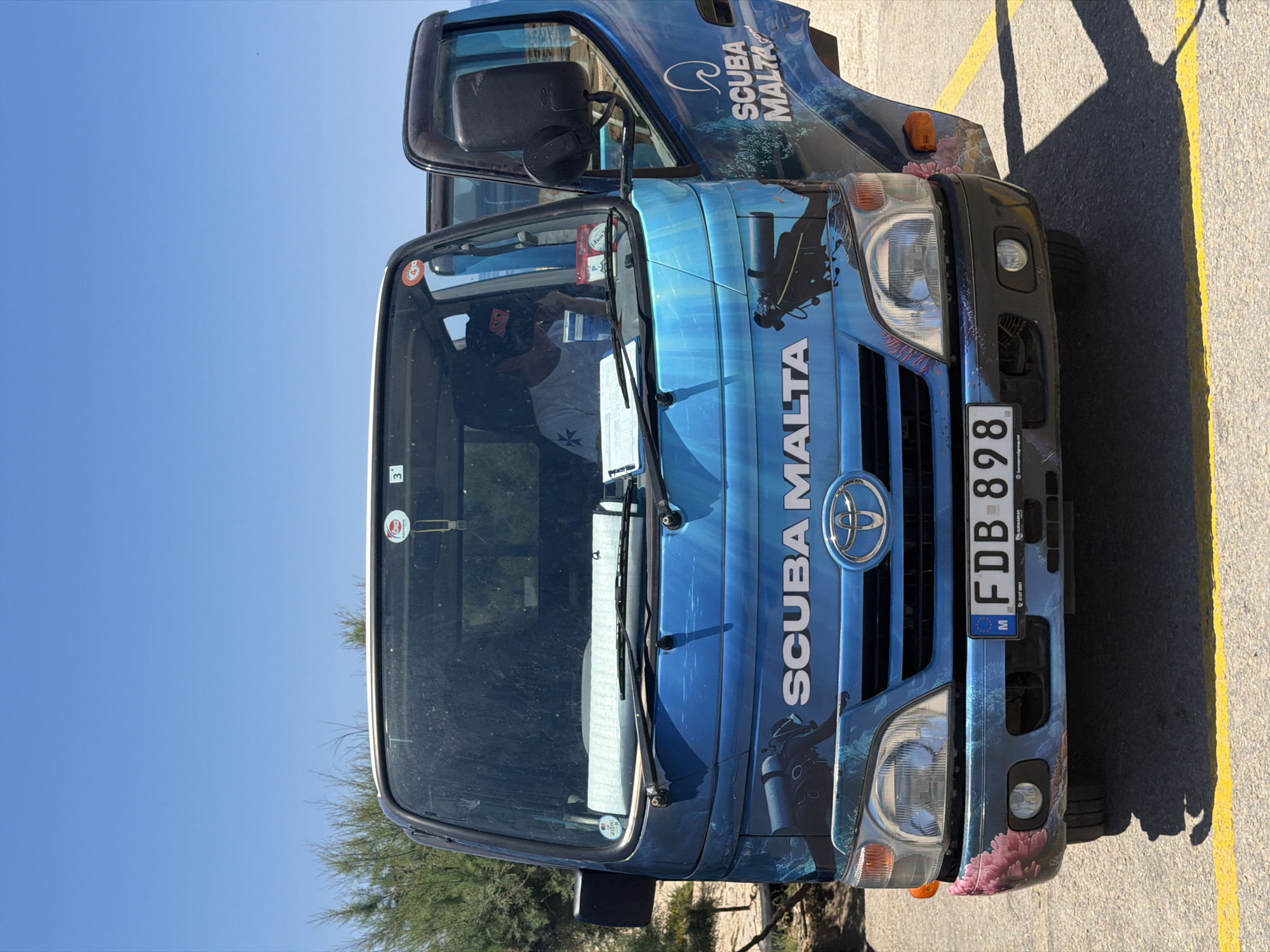 A blue Toyota truck with a scuba diving theme is parked on an asphalt surface marked with yellow lines. The truck's design features underwater imagery, including coral, fish, and a diver. The words "SCUBA MALTA" are prominently displayed on the front and side of the vehicle. The truck's license plate reads "FDB 898" and indicates Malta. A person wearing a hat and white t-shirt is visible inside the truck's cabin, appearing to be engaged with a clipboard. The background shows a clear blue sky and some sparse vegetation, consistent with a coastal or arid environment. The image appears to be taken during the daytime under bright sunlight, casting shadows on the ground. The location is Saint Lawrence, Malta.