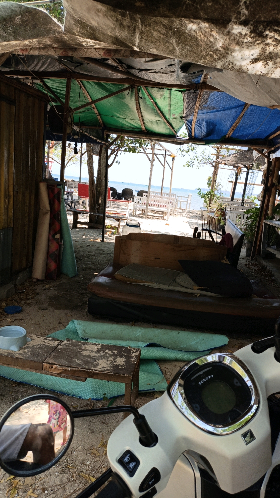 The image depicts an outdoor scene, likely in Cikal, Indonesia, showcasing a beachside area with a makeshift shelter. The shelter, constructed with corrugated metal walls and a canopy made of various tarpaulins in green, blue, and grey, covers a seating area and storage space. In the foreground, a white scooter is parked, its digital display visible, reading "SCOOPY." To the left, a side mirror reflects a partial view of a person wearing a pink dress and a person's legs.
Dominating the mid-ground is a large, worn sofa with cushions. Rolled-up mats are stacked against the left wall, and another rolled-up mat lies on the sandy ground. A rustic wooden table with a blue bowl on it sits near the mats.
The background reveals a sandy beach with several white wooden benches and chairs, suggesting a seating area for patrons or visitors. Beyond the seating area, the calm sea is visible under a clear sky. Trees with sparse foliage are scattered throughout the scene, and some potted plants are also present. The overall impression is of a casual, possibly under-renovation, beachside establishment.