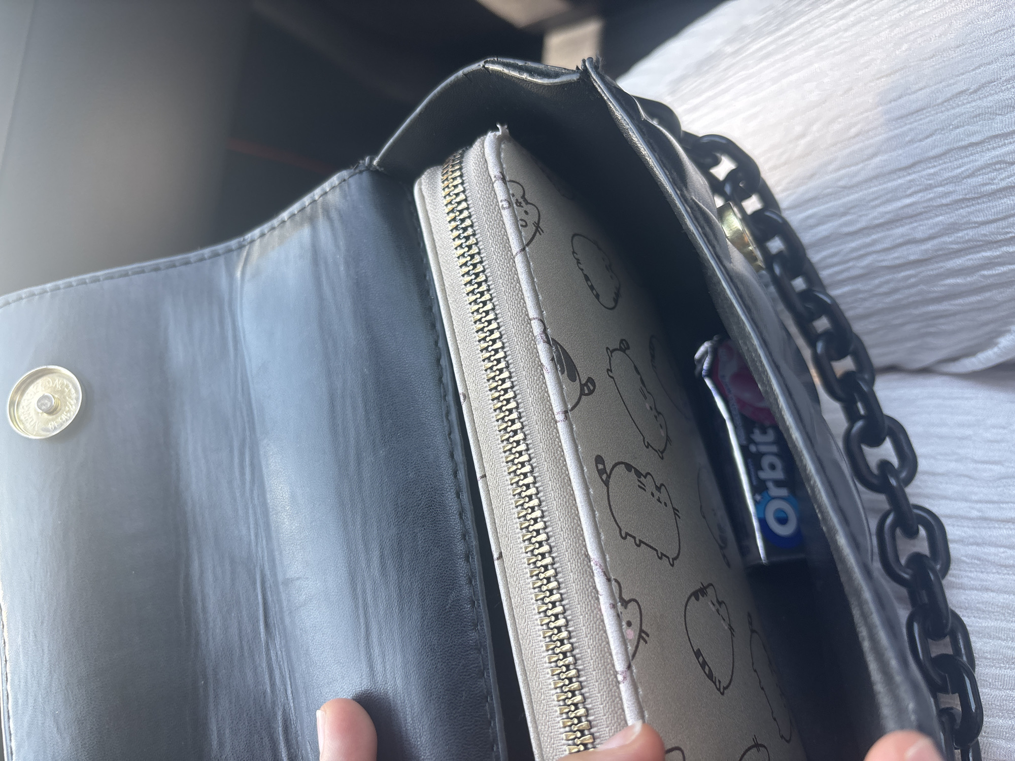 An overhead, close-up shot shows the contents of a black purse. A person's thumb and forefinger are visible at the top left, holding open the purse's flap. Inside, a light-colored wallet with a zipper closure is partially visible. The wallet has a pattern of cartoonish cats. Resting on top of the wallet is a pack of Orbit chewing gum. The purse has a chunky black chain strap. The background is out of focus, but suggests an indoor setting, possibly a car interior. There is no visible indication of the city of Sofia, Bulgaria.
