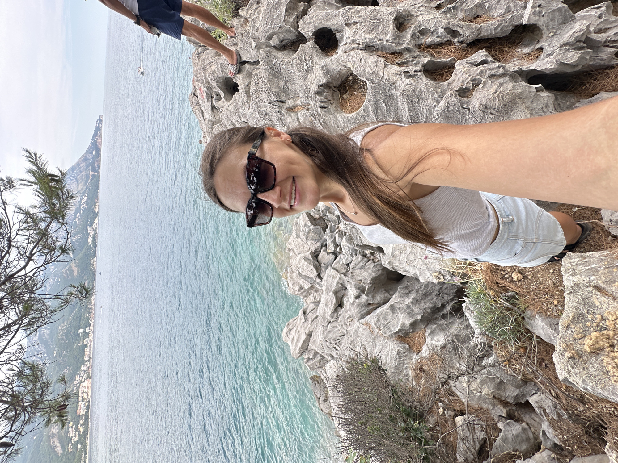 A woman with long brown hair and sunglasses is taking a selfie while standing on a rocky cliff overlooking the Adriatic Sea in Budva, Montenegro. She is wearing a light grey tank top and denim shorts. The sea is a beautiful turquoise color, and a coastline with buildings and lush greenery is visible in the distance. The sky is bright, suggesting it is daytime with good weather. There is another person in the background, partially visible, wearing blue shorts and standing on the rocks. The terrain is rugged and rocky, with some dry grass and pine needles scattered on the ground. The woman is smiling, appearing happy and enjoying the scenic view.