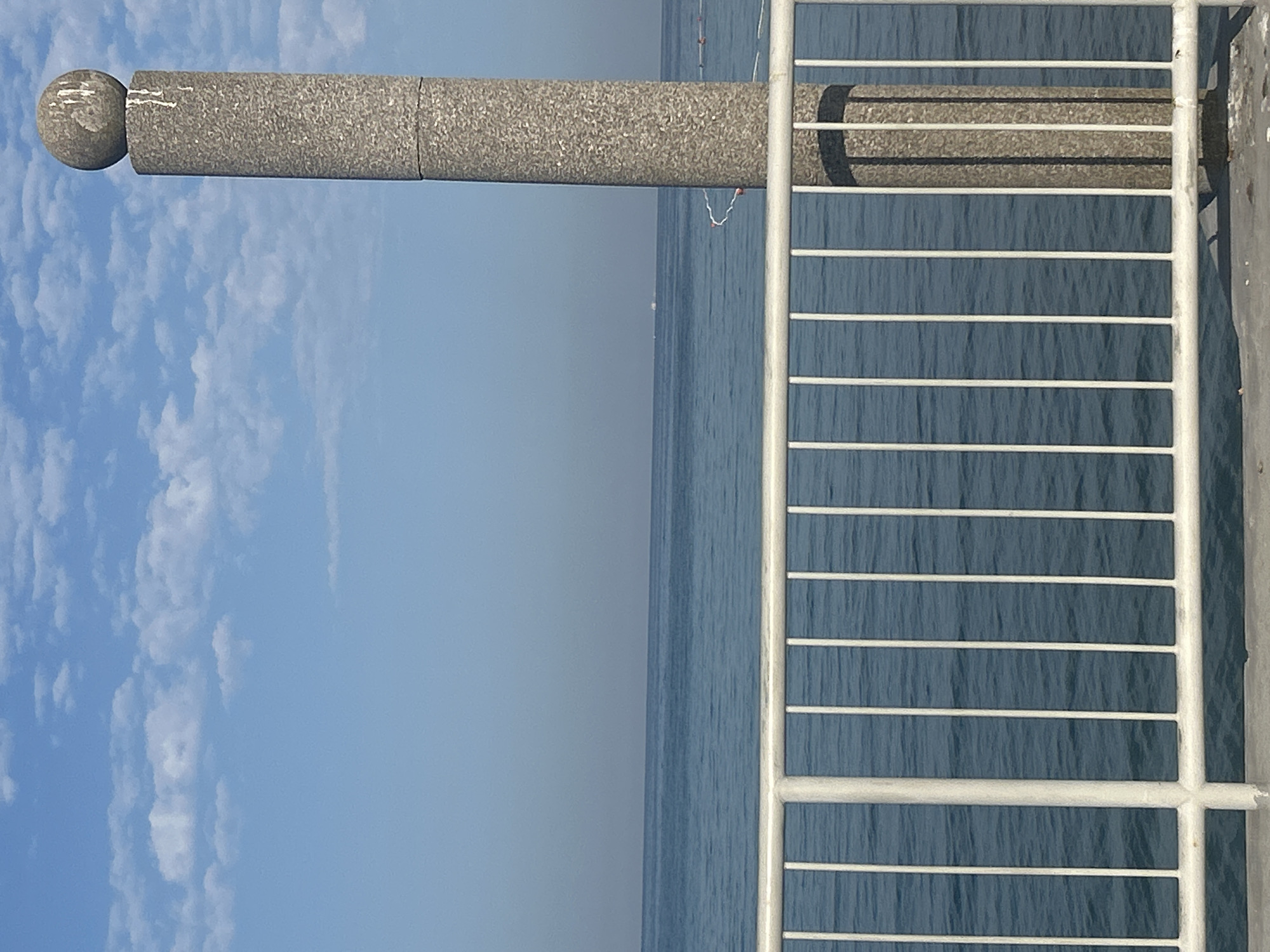 The image depicts a clear day with a blue sky and scattered white clouds. In the foreground, a white metal railing runs horizontally across the frame, with vertical bars spaced evenly. Behind the railing is a body of calm blue water, likely the sea. In the right half of the image, a tall, cylindrical concrete pillar stands, topped with a spherical stone element. The pillar has a textured, speckled surface. In the distance, on the horizon where the water meets the sky, faint white specks suggest distant boats or landforms. The overall scene evokes a sense of tranquility and open space. The description provided does not contain specific location details of Monterol, Croatia.