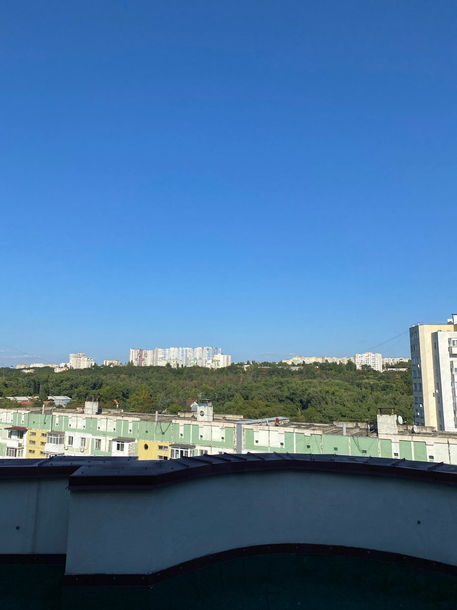 The image shows a panoramic view of a residential area in Chișinău, Moldova, under a clear blue sky. In the foreground, there is the dark, curved edge of a rooftop with a maroon trim. Behind this, a row of buildings with green and yellow facades are visible. These buildings appear to be apartment blocks, with multiple windows and balconies. Beyond these, a dense, lush green forest covers a hillside. In the distance, several large, multi-story apartment buildings with white and light-colored facades rise above the trees, indicating the presence of a more densely populated urban area. The architecture of these distant buildings suggests a Soviet-era or post-Soviet style. The scene is bathed in sunlight, casting shadows and highlighting the details of the buildings and the vegetation. There are no people or vehicles visible in the image, and the overall impression is one of a quiet, daytime urban landscape.