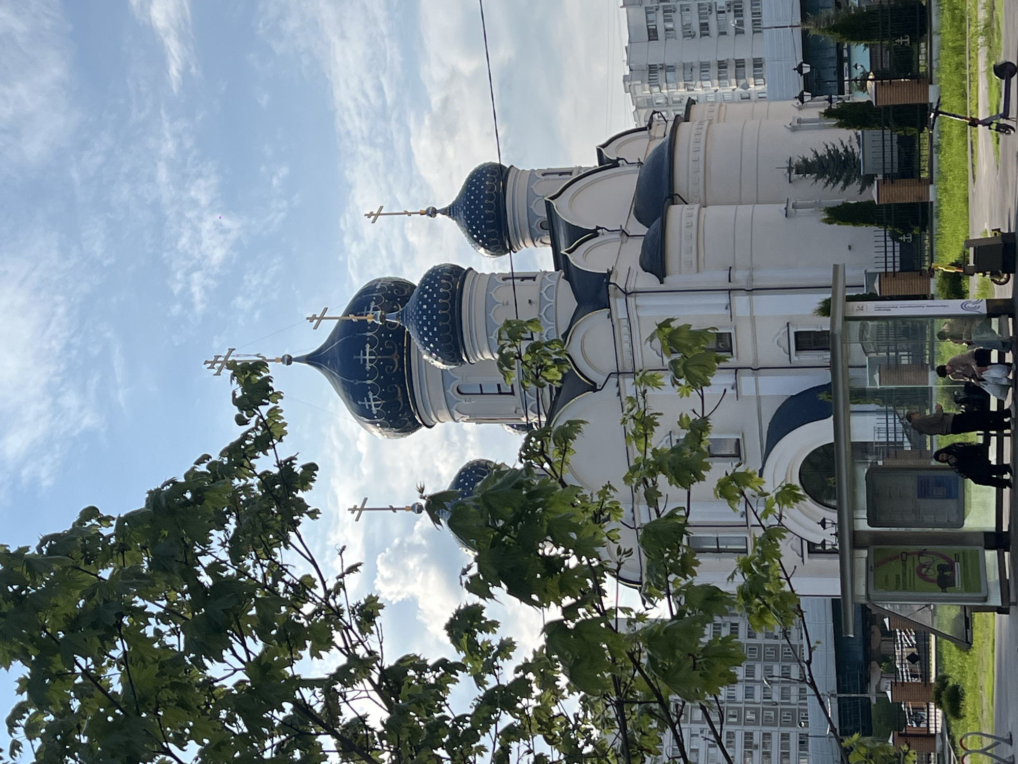 The image shows the Donskoy Monastery in Moscow, Russia. The monastery is seen from a low angle, partially obscured by a tree with green leaves. The architectural details of the monastery are evident, including its white walls and blue domes topped with golden crosses. In the background, a modern residential building is visible. A bus stop is in the foreground, where people are waiting. The sky is blue with white clouds.