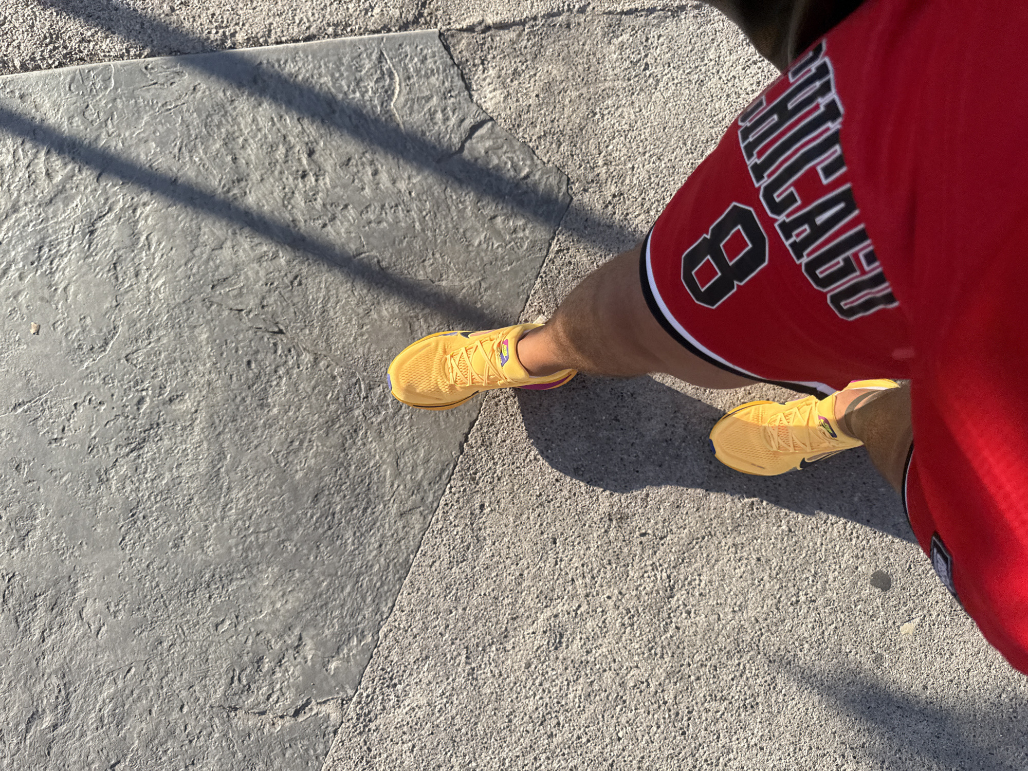 A low-angle, outdoor, point-of-view shot shows a person walking on a textured concrete surface. The person is wearing bright yellow athletic shoes with purple and pink accents, red basketball shorts with the number "8" and the word "CHICAGO" in black and white lettering, and a brown object, possibly a bag or jacket, is visible on the right side of the frame. The ground is composed of two different textures of concrete, with shadows cast across the surface, suggesting a sunny day. The scene is set on a pedestrian walkway, likely in a public area like a promenade or park. There is no visible indication of the specific location in Budva, Montenegro.