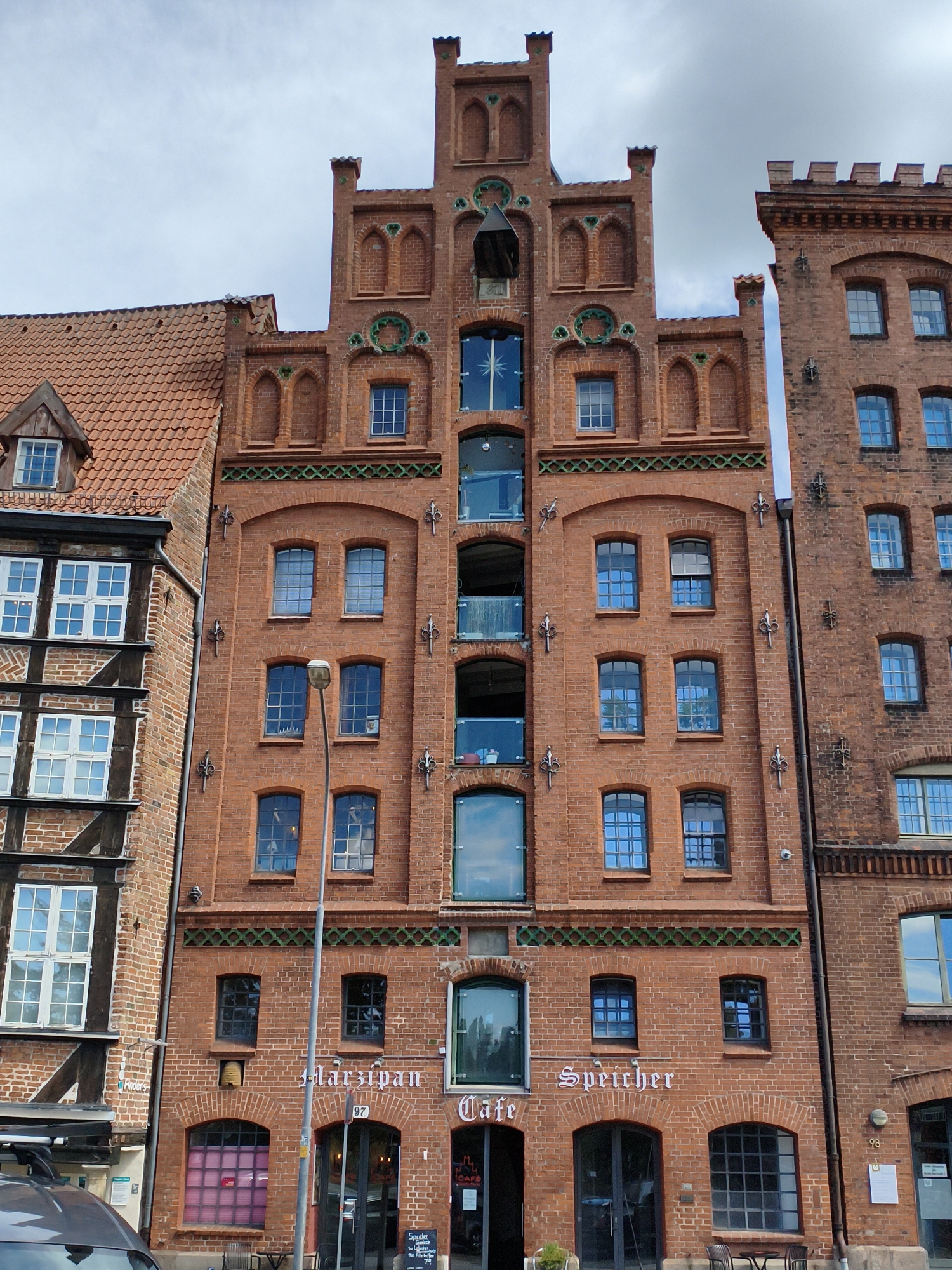 The image shows a row of brick buildings in Lübeck, Germany. The building in the center has a sign that reads "Marzipan Cafe Speicher". The architecture features ornate brickwork, arched windows, and pitched roofs. The sky is overcast. In the foreground, a portion of a car is visible. There are no people clearly visible in the image. The buildings appear to be old, possibly historic warehouses converted into shops or cafes.