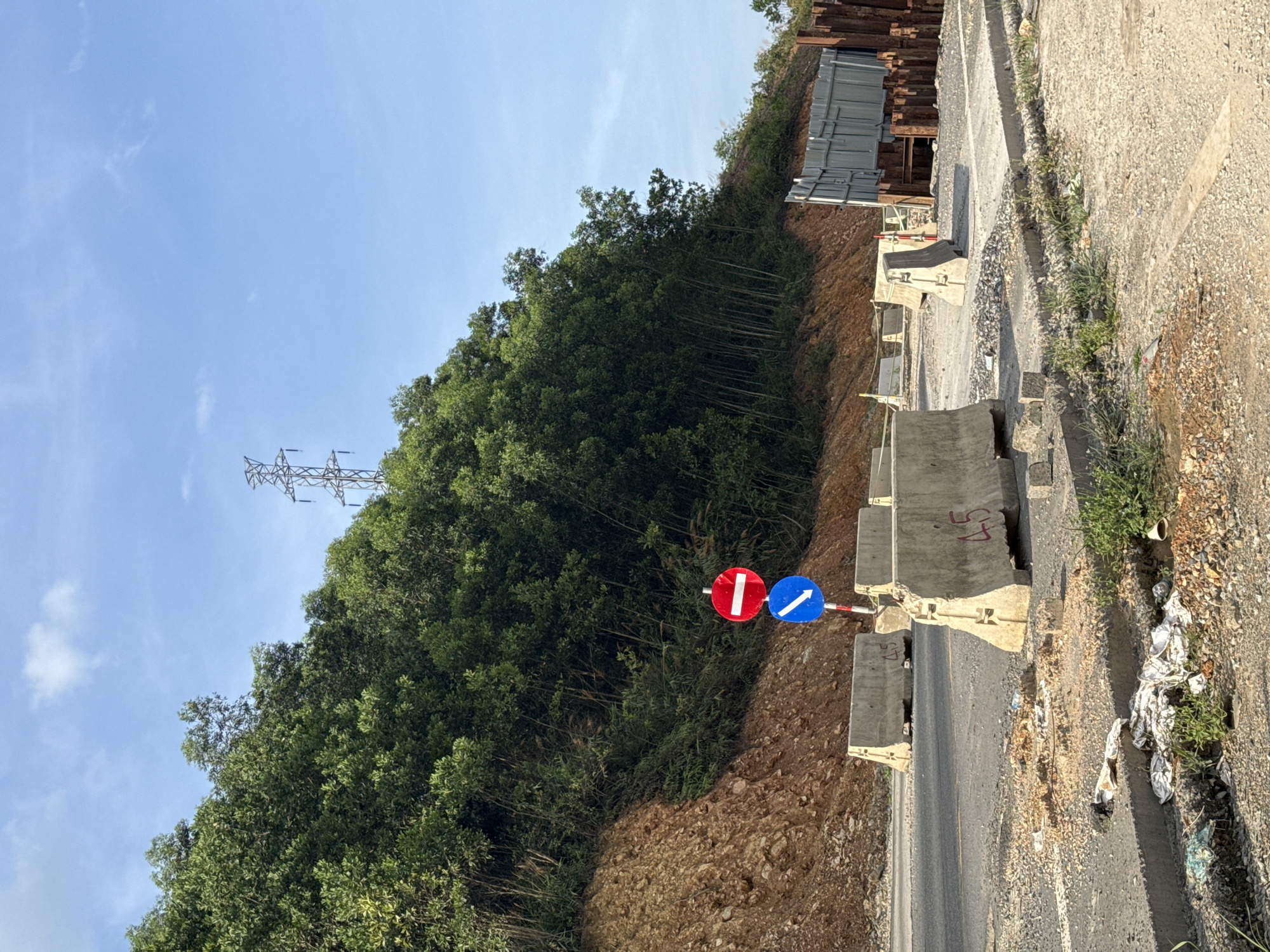 The image shows a road construction scene near a hillside in Đà Nẵng, Vietnam. A roadside ditch is filled with trash. Concrete barriers line the edge of the road, with "45" spray-painted on one. Two road signs are visible: one red circle with a white horizontal line (no entry), and another blue circle with a white arrow pointing to the right (mandatory direction). A lush green hillside rises on the left, with utility poles and power lines visible in the background. The sky is partly cloudy, suggesting daylight conditions. Construction materials, like steel beams, are stacked behind the barriers further up the hill. The road itself is asphalt, and it appears to be an active construction site.