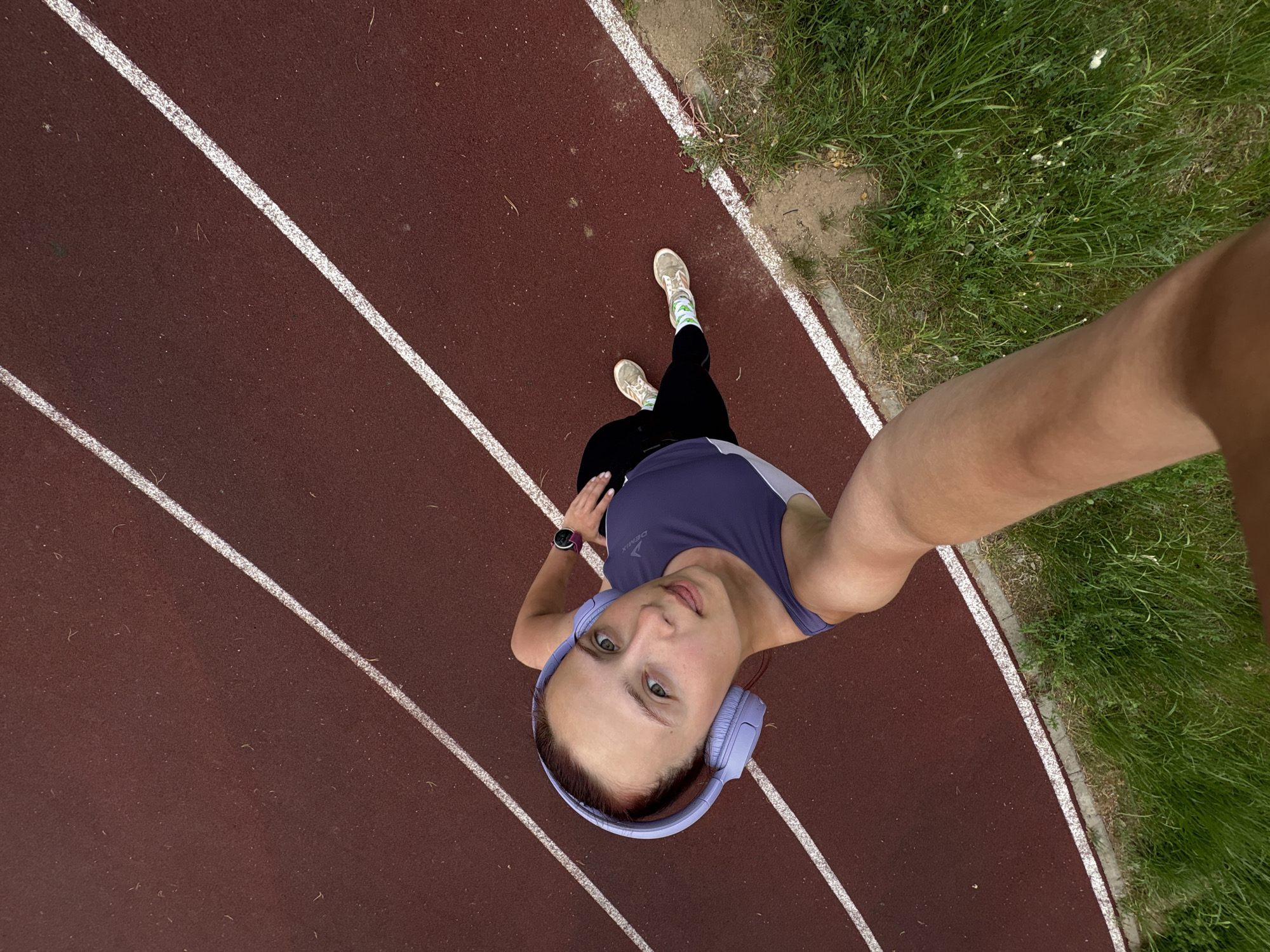 A woman is taking a selfie on a running track in Прикамский, Russia. She is wearing a purple tank top, black pants, and sneakers. She has headphones on and is wearing a watch. The running track is red with white lines. There is grass on the side of the track.