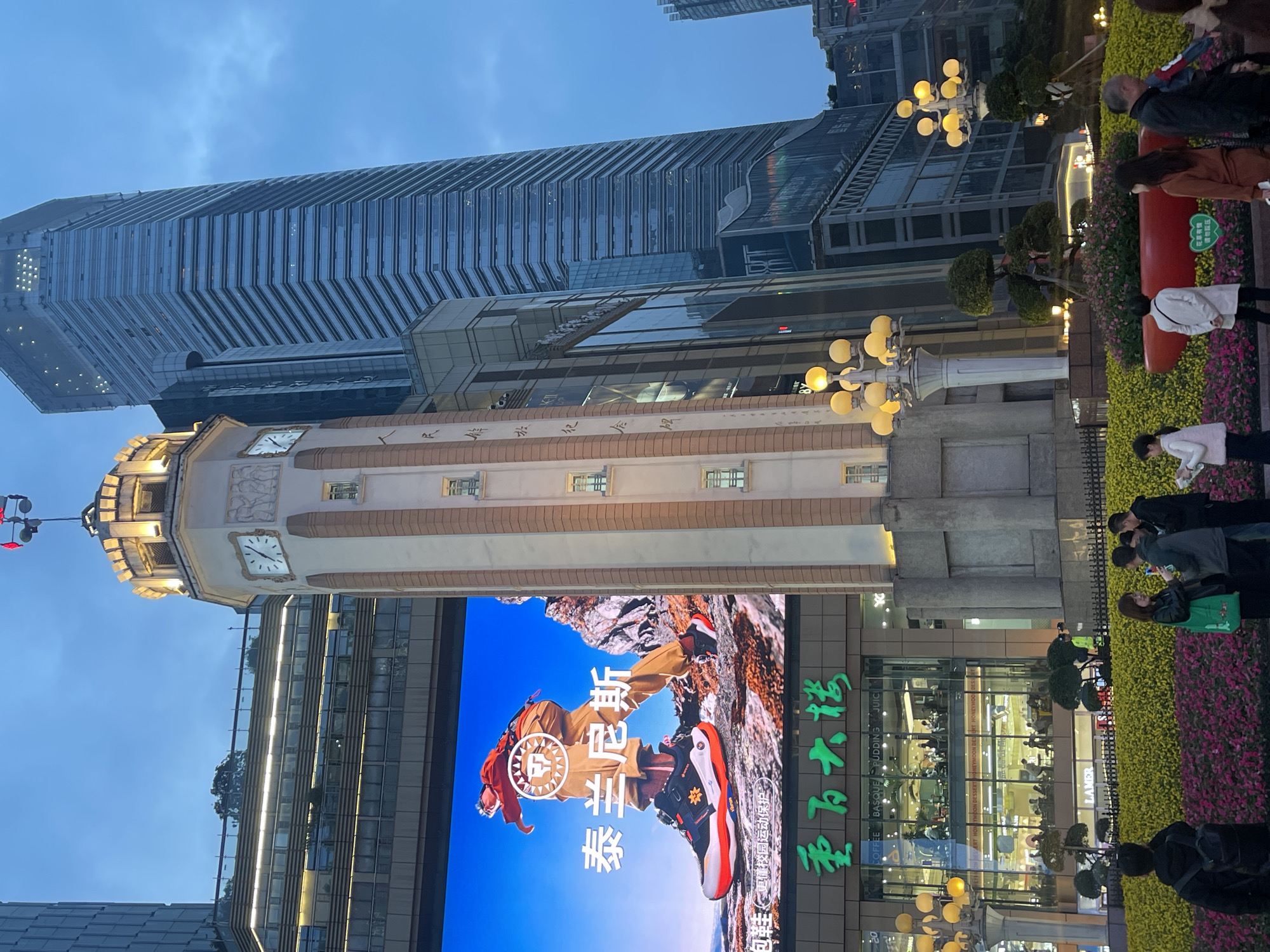 The image depicts an urban scene at dusk or early evening on Jiefangbei Pedestrian Street, Yuzhong District, Chongqing, China. In the center, a multi-story, cream-colored clock tower with brown vertical accents stands prominently. It features illuminated clock faces on at least three visible sides and a finial with red flags on top. Vertical Chinese characters are inscribed on its body.
To the left of the clock tower, a large commercial building displays a massive illuminated LED screen showcasing an advertisement for "Tersan" (泰兰尼斯) athletic shoes, featuring a person in hiking gear on a rocky terrain. Below the LED screen, the green neon sign "重百大楼" (Chongqing Department Store) is visible, with lower levels featuring glass windows revealing interior restaurant or retail spaces populated by numerous individuals.
To the right and behind the clock tower, a tall, modern skyscraper with a distinctive corrugated glass facade rises into the darkening sky, its upper sections reflecting the blue sky. Additional urban structures are visible in the background.
In the foreground, a landscaped pedestrian area features extensive flowerbeds filled with yellow and pink/purple blossoms, interspersed with smaller ornamental trees in planters. Multiple ornate, multi-bulb streetlights with warm yellow illumination are active. Numerous pedestrians are present in the midground and foreground, walking or standing in small groups. Individuals are dressed in casual attire, some appearing to converse or look at phones. One person in the immediate foreground wears a black hoodie and backpack, facing away from the viewer.