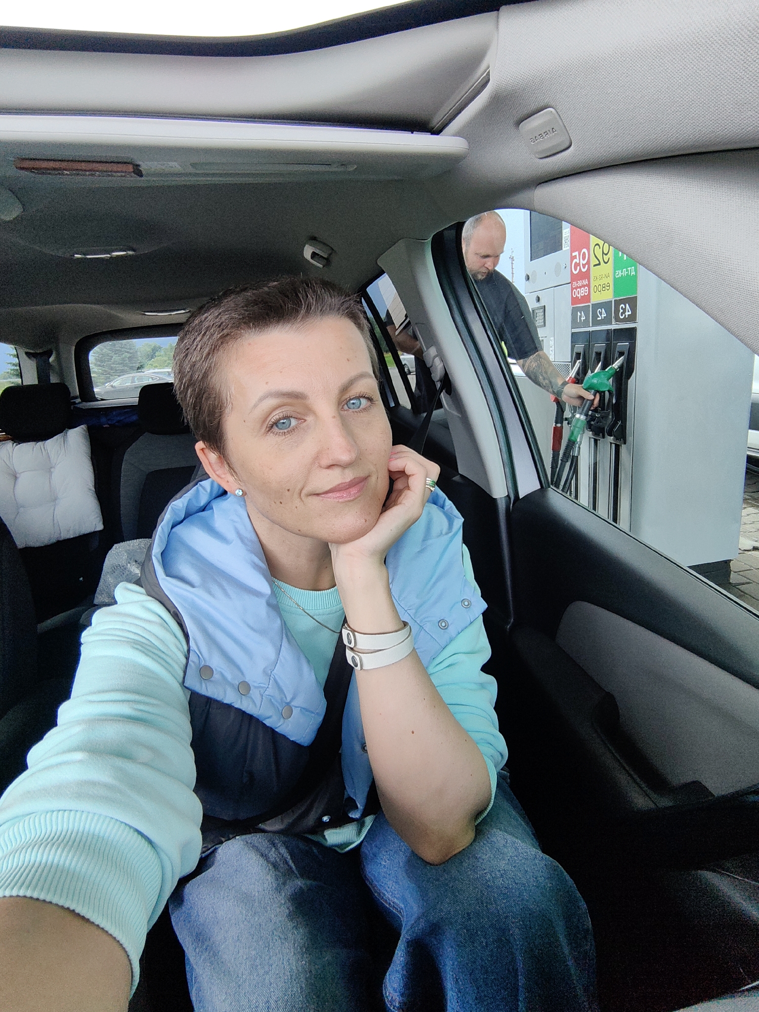 A woman with short brown hair and blue eyes is sitting in the passenger seat of a car at a gas station. She is wearing a light blue hooded vest over a light blue sweatshirt and blue jeans. Her right hand is resting on her chin. In the background, a bald man with tattoos on his arm is visible, holding a green gas nozzle at a fuel pump. The gas station signage displays prices in Belarusian rubles. The scene appears to be the gas station located on the M1 highway near the city of Fanipal, in the Fanipal rural council of Belarus.