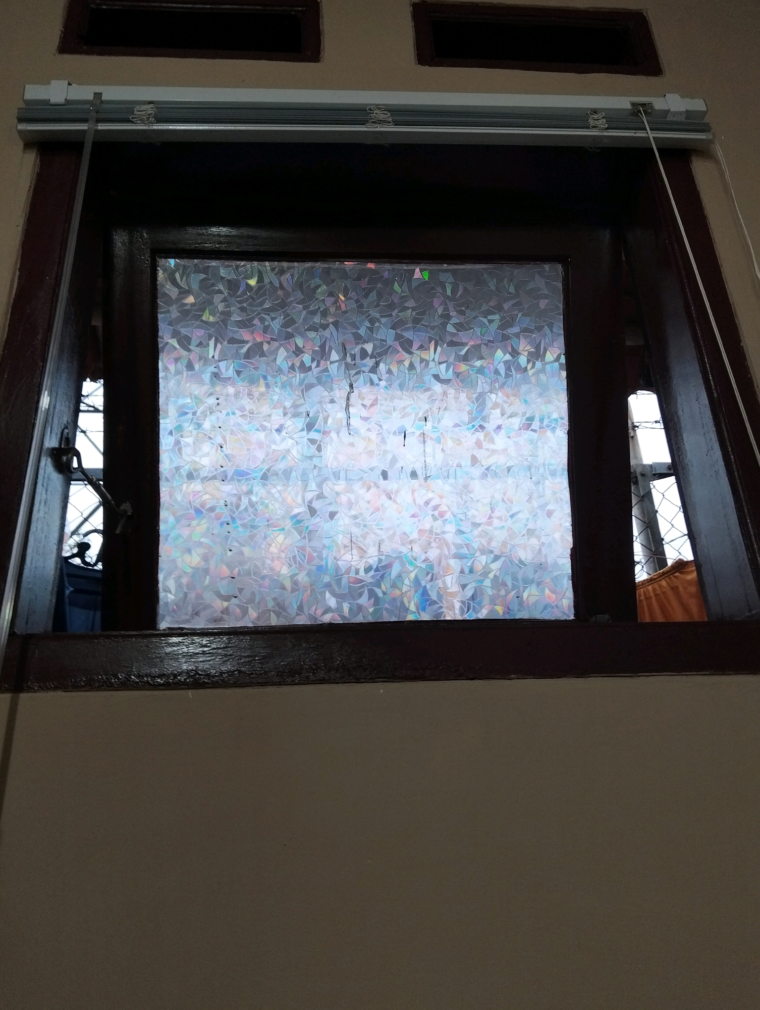 The image shows a window with a decorative film that refracts light into a mosaic of colors. The window is framed by dark wood and is part of a building with beige walls. A white roller blind is mounted above the window. The window is partially open, revealing a glimpse of the exterior, which includes a metal grate and an orange fabric. The scene is likely an interior shot of a residential or commercial building in Patrol, Indonesia.