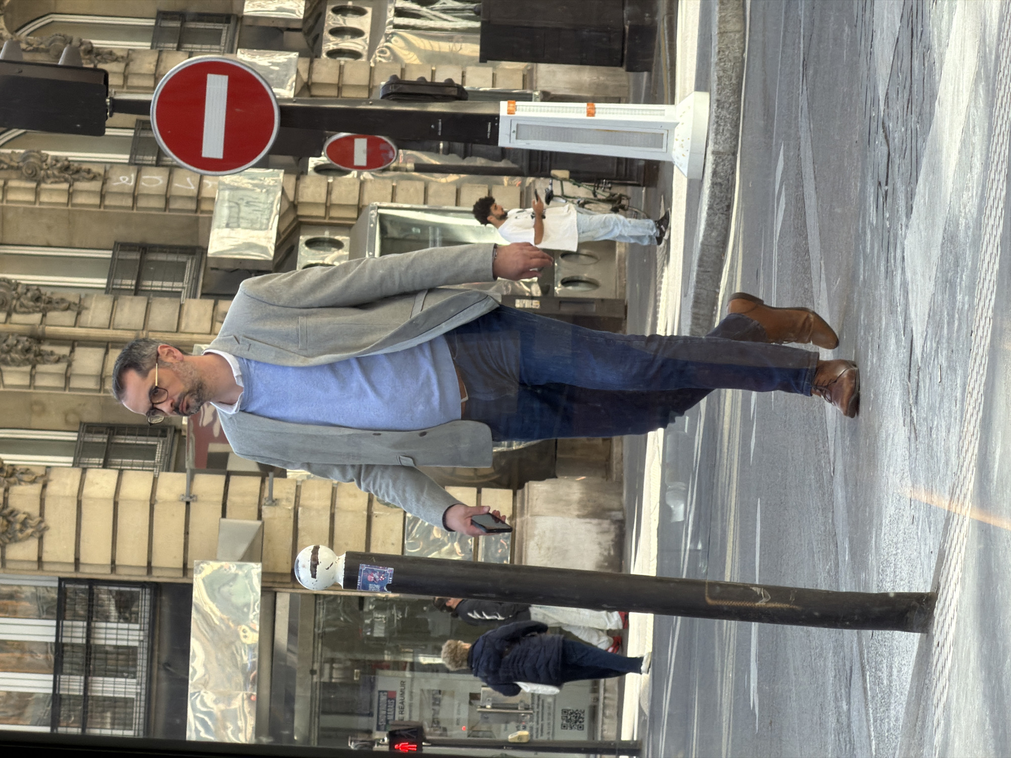 The image appears to have been taken in Paris, France, judging by the architectural style visible in the background. It focuses on a man dressed in a light blue sweater, a grey blazer, and dark jeans, walking while looking at a smartphone in his hand. There are street signs in the background, and other people walking or standing nearby, including one person in a white uniform. The street surface is light-colored, possibly concrete or asphalt, and seems to reflect sunlight.