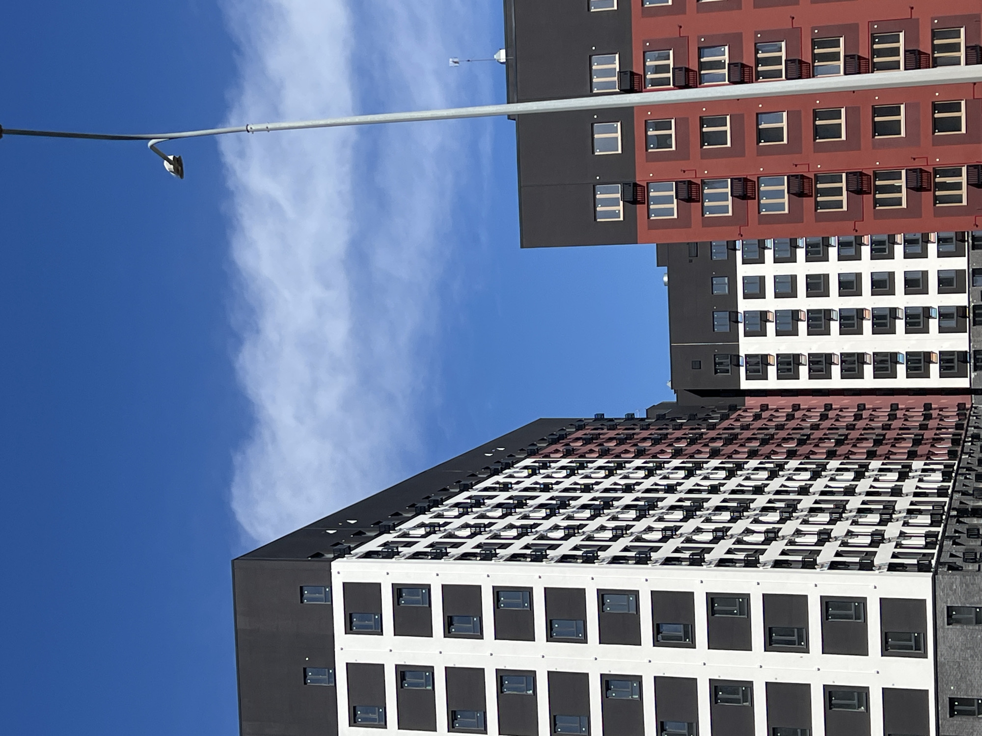 The image shows a view of several modern apartment buildings under a blue sky with scattered clouds. A street lamp pole stretches across the upper left of the frame. The buildings are characterized by a grid-like facade, with alternating colors like white, black, and red. Each module of the grid contains windows and small protruding structures that might be air conditioning units or decorative elements. The architecture of the buildings is modern and uniform. The image is taken at an angle, tilting the scene. The buildings are located in Tyumen, Russia.
