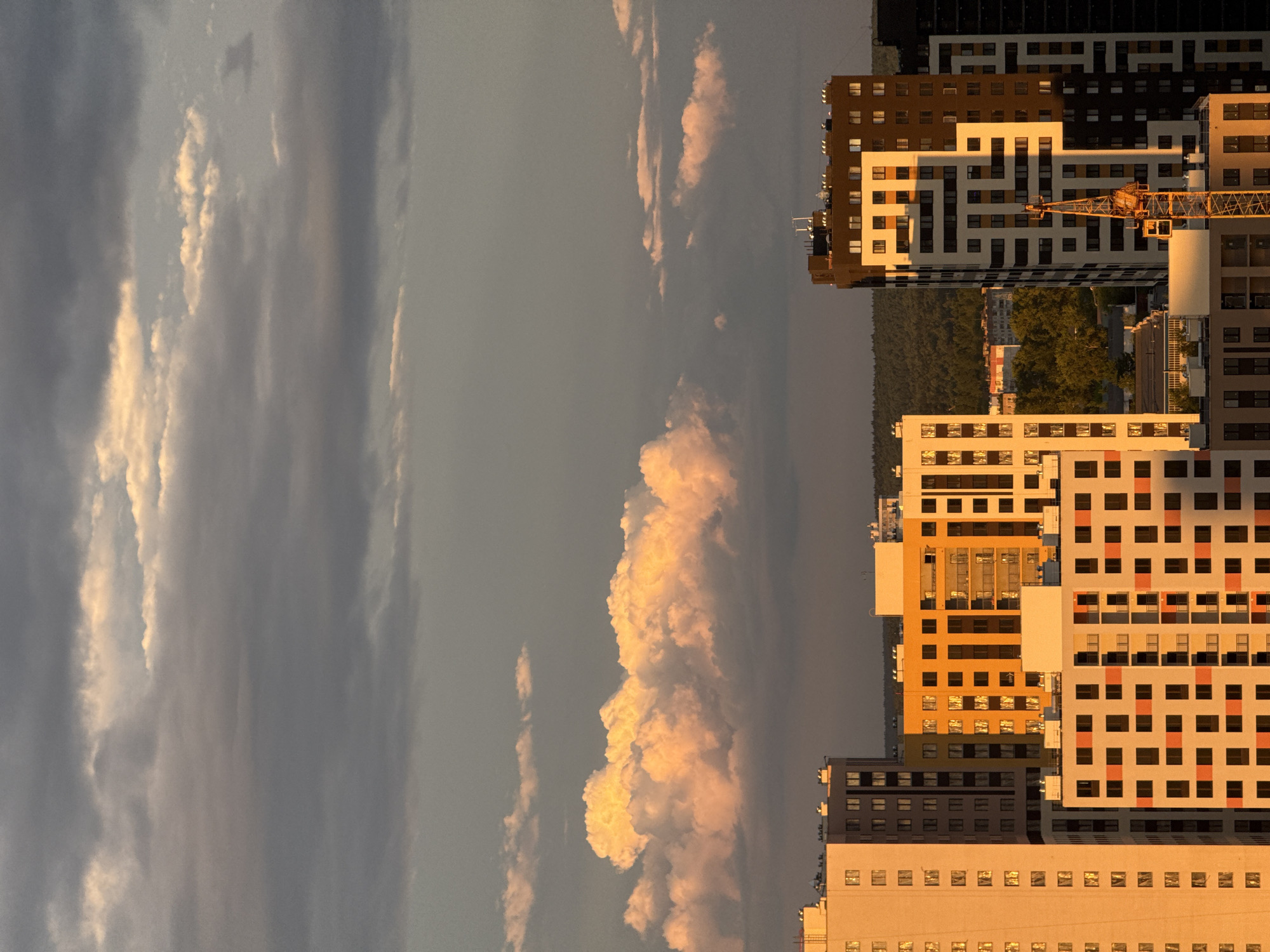 The image depicts a cityscape with buildings on the right and a dramatic sky on the left. The buildings are multi-story, with a mix of light and dark colors, including white, orange, and brown. A construction crane is visible among the buildings. The sky is filled with clouds, some of which are brightly lit, suggesting either sunrise or sunset. The location appears to be in a city, with a mix of residential and possibly commercial buildings. The trees are green, which suggests the setting is a relatively temperate or warm climate. The overall tone is serene, with the soft light creating a sense of calm.