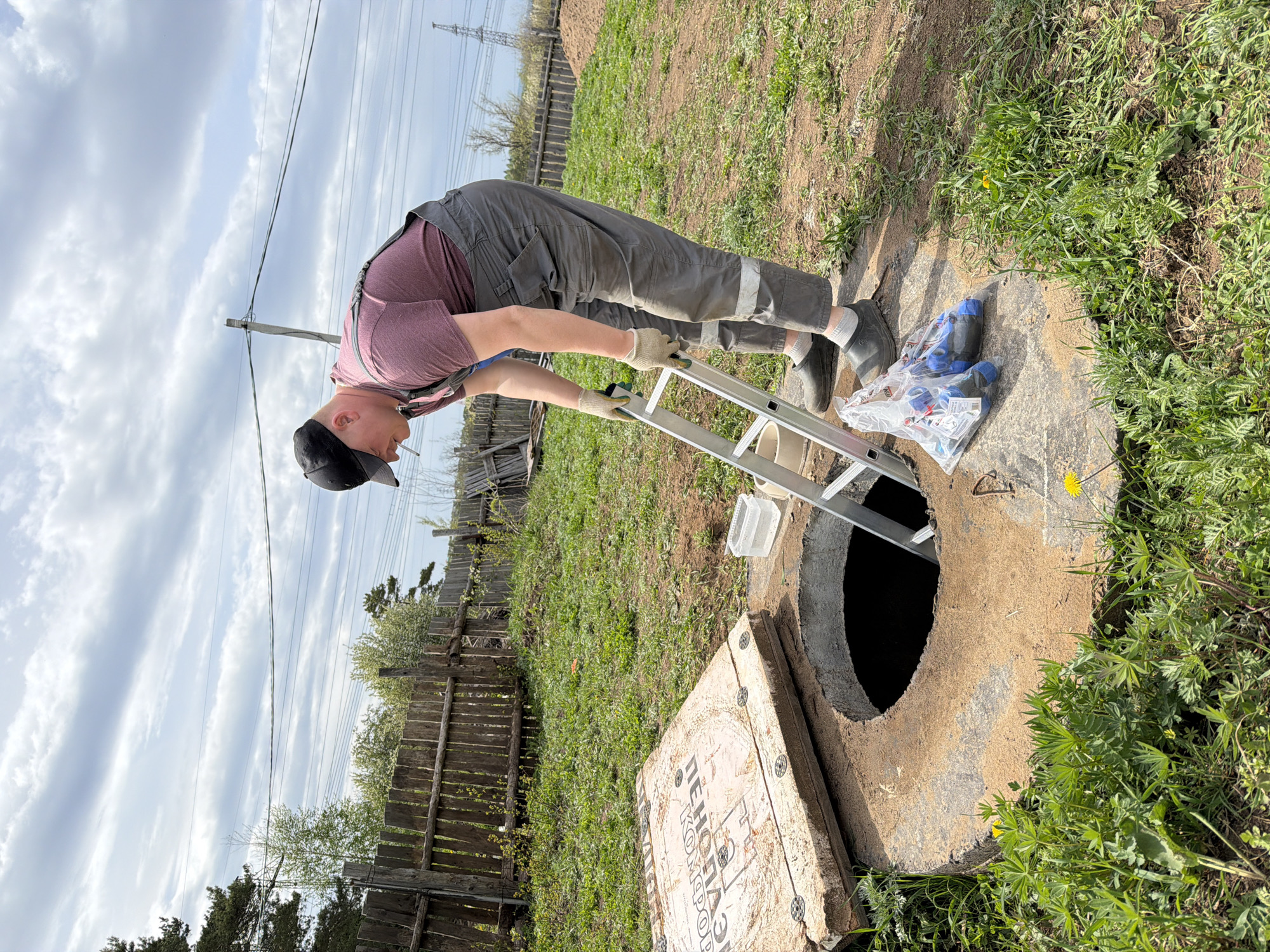 A man in work attire is setting up a ladder to descend into a concrete manhole. He is wearing gray overalls, a maroon shirt, gloves, and a black cap. A cigarette is in his mouth. The manhole cover is displaced next to the opening. A bag of blue shoe covers and a small white container are near the manhole. The surrounding area is grassy, with a wooden fence in the background. Overhead power lines are visible. The location is Chaikovsky, Russia.