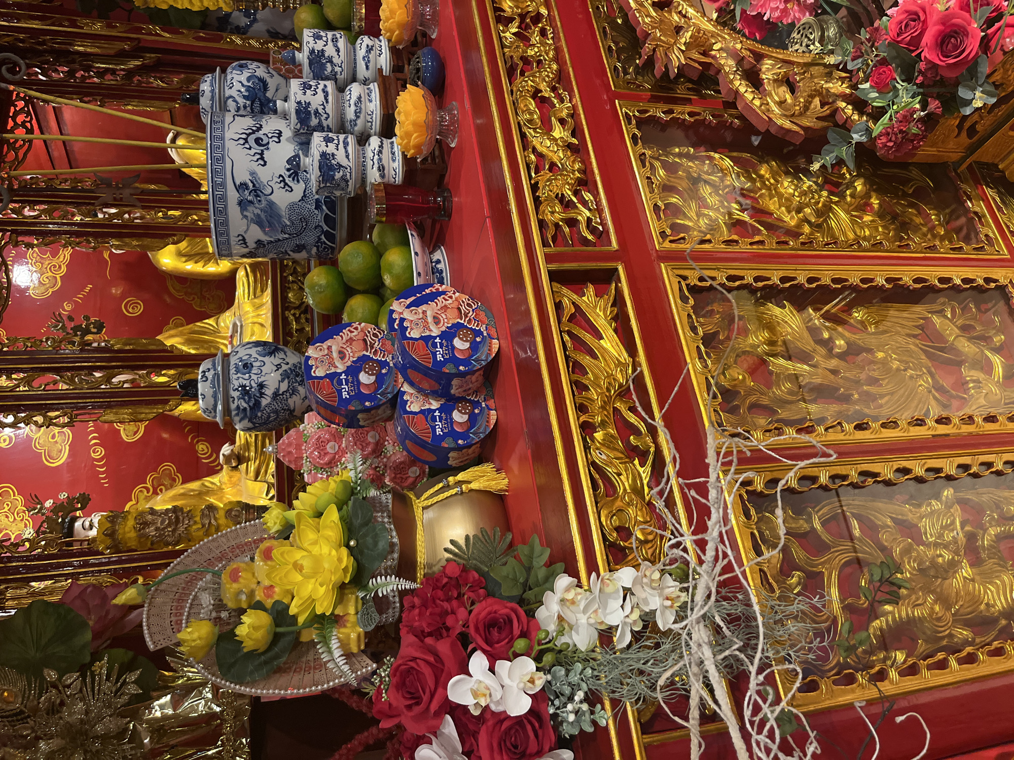 The image depicts a heavily decorated altar in a location in Hà Nội, Vietnam. The altar is adorned with various items: stacks of blue and white porcelain containers, possibly incense holders; several stacks of blue circular containers with Japanese writing on the labels; arrangements of yellow and red flowers; small bowls containing liquids and fruit; and red and gold architectural elements that appear to be part of the altar's construction. Golden dragon carvings are prominent. The lighting appears to be indirect. The image captures a scene of religious or cultural significance within a Vietnamese context.