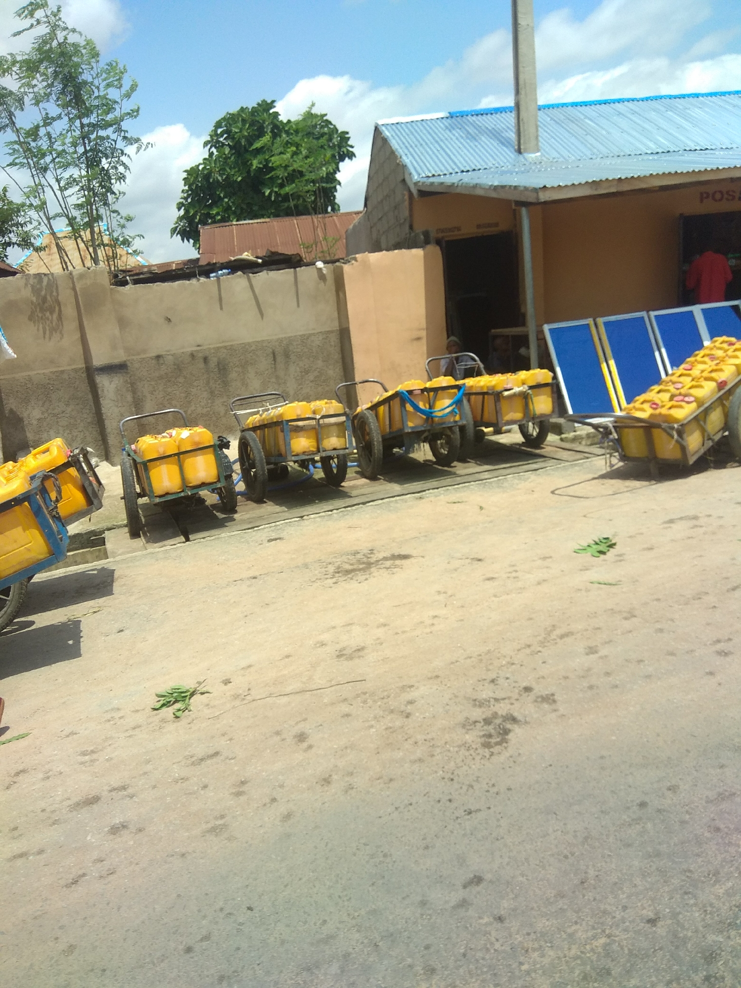 In Jalingo, Nigeria, a row of yellow plastic jerrycans filled with liquid are loaded onto several carts, which are lined up on a dirt road in front of a building with a corrugated metal roof. The carts, some with metal frames and others with blue and yellow frames, are designed to transport the jerrycans. The scene appears to be a marketplace or a point of sale. The sun is bright, indicating it is daytime, and the sky is partly cloudy. The building in the background has a light brown facade and appears to be a shop or business. The word "POSA" is visible on the building's facade. The overall atmosphere is one of commerce and daily activity. There are some plants and debris scattered on the road, suggesting a typical outdoor setting. The image captures a moment of logistical activity, likely related to the distribution or sale of the contents of the jerrycans.