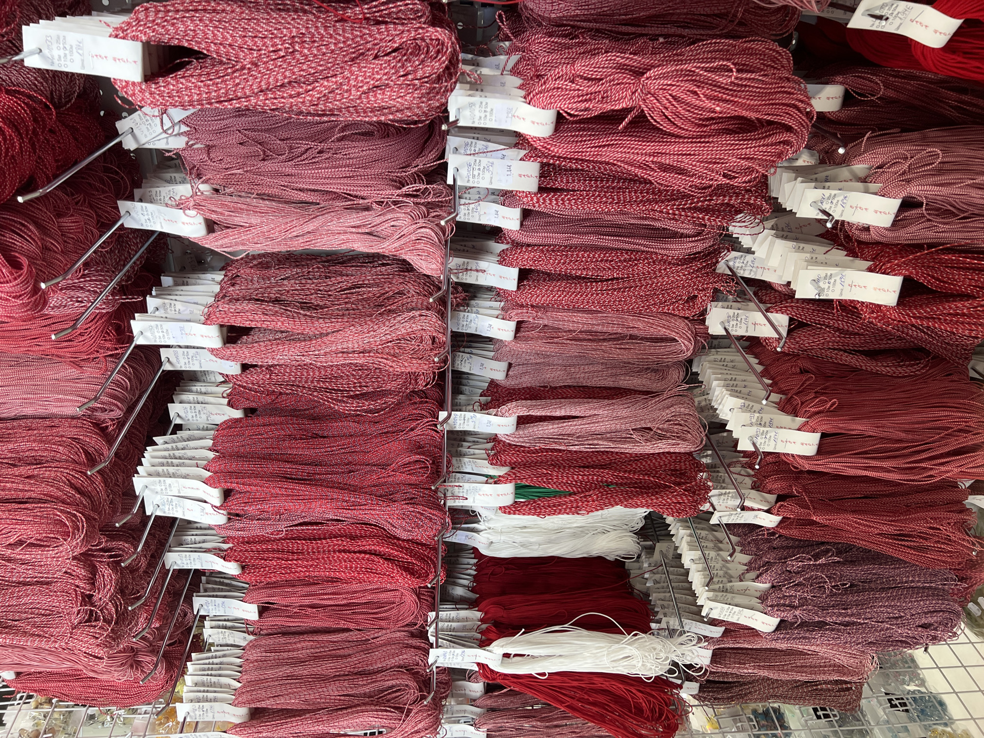 The image shows a close-up view of shelves stocked with bundles of yarn, primarily in various shades of red, pink, and white. Each bundle appears to be tied with a small tag, possibly containing information about the yarn. The shelves are made of metal, with thin rods supporting the merchandise. The yarns are neatly organized, suggesting a retail setting where craft supplies are sold. The scene appears to be indoors.