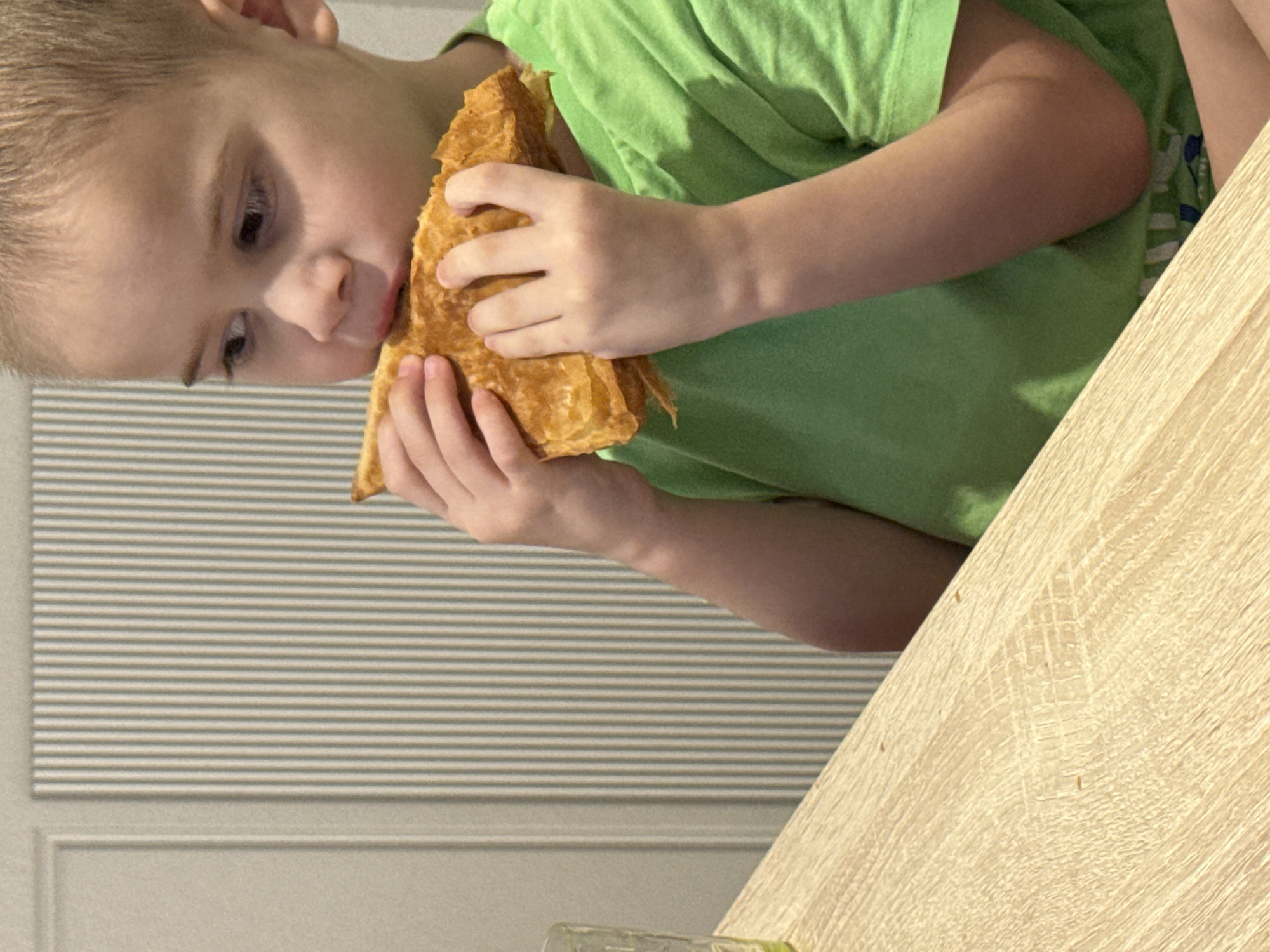 A young, light-skinned male child with short, light brown hair, wearing a bright green short-sleeved t-shirt, is depicted indoors at a table in Chaikovsky, Russia. He is positioned on the right side of the frame, facing slightly left but with his gaze directed towards the right. His hands hold a golden-brown, layered pastry or flatbread to his mouth, with his upper lip pressed against the food, indicating active consumption. A portion of a light-colored, textured wooden table is visible in the lower-left quadrant, where a clear, textured glass is partially present. The background features a light-colored wall with distinct vertical paneling; the left section displays a smooth panel with a recessed vertical line, transitioning to vertically ridged or fluted paneling across the central and right portions of the wall.