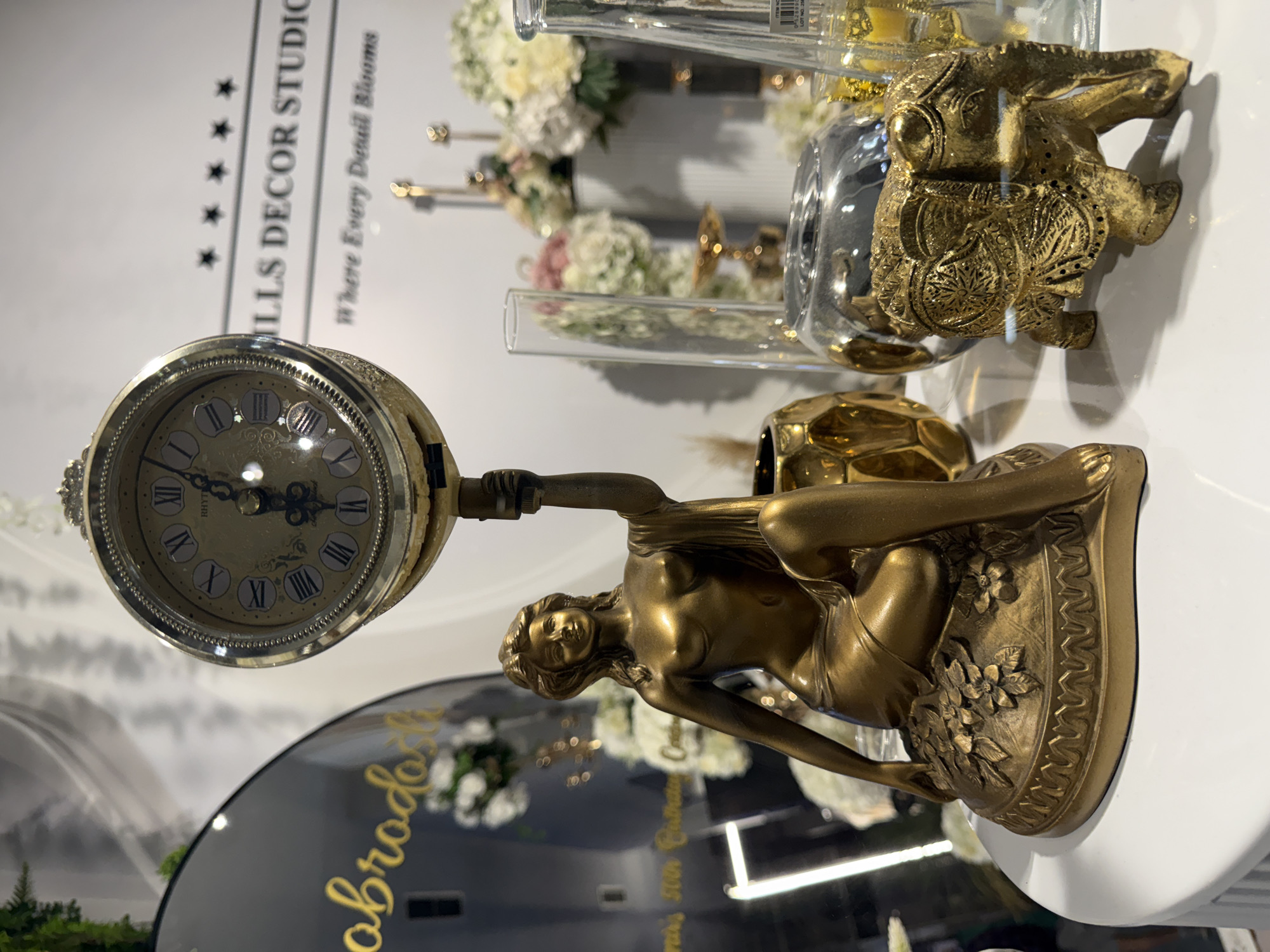 This image depicts a retail display in Ilidža, Bosnia and Herzegovina, showcasing various decorative items. The foreground features a golden figurine of a woman holding a clock aloft, seated on a decorative base. To the right of the figurine, a small golden elephant statue is visible, with intricate carvings. In the background, floral arrangements in vases, glass cylindrical vases, and a large black circular mirror with text are present. A sign with the text "MILLS DECOR STUDIO Where Every Detail Blooms" is also discernible in the background. The overall setting suggests an interior shop environment, likely a home decor store, with items displayed on a white surface.