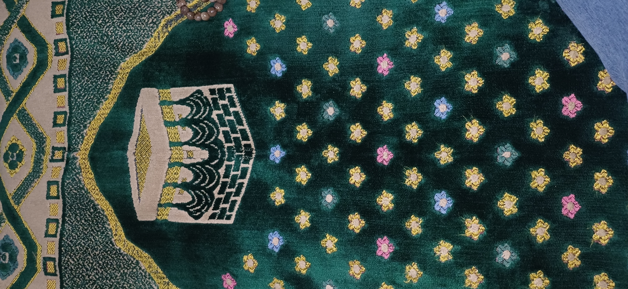 A close-up, top-down view shows a plush, dark green prayer rug with a repeating pattern of small, multi-colored floral motifs and a larger, central design of the Kaaba. The Kaaba is depicted in beige and green, with gold trim, and appears to be framed by a textured green fabric. A string of prayer beads, light brown and grey, is draped along the top right edge of the rug. The background of the rug features a patterned geometric design in beige and gold. The rug is positioned on an unseen surface, with a sliver of blue fabric visible at the bottom right corner. The scene is indoors, with no discernible city or environmental context.