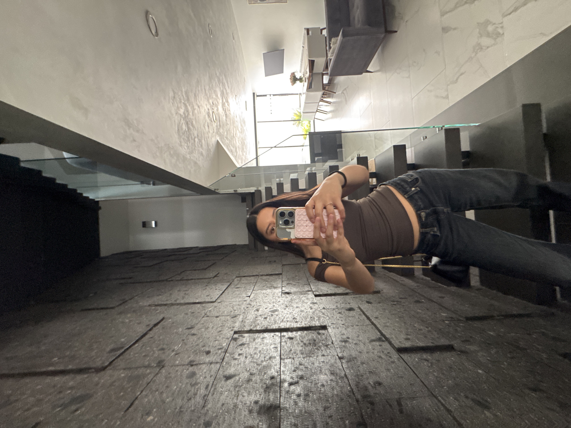 A young woman with long brown hair is taking a selfie in what appears to be a modern, upscale residence. She is wearing a brown top and dark jeans, holding a smartphone with a pink case. The room has a gray tiled floor and a glass staircase railing. In the background, there is a glimpse of a dining area with a long table and chairs, as well as a living room with a gray sofa. The location is possibly in Puebla, Mexico.