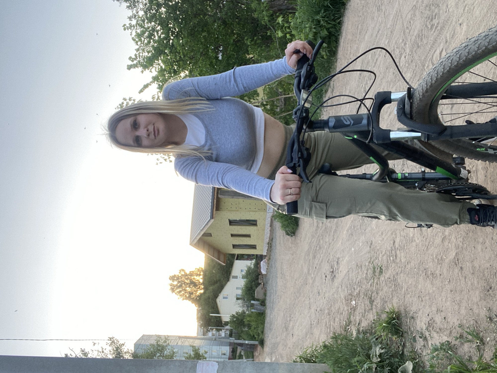 A young woman with blonde hair is posing on a mountain bike. She is wearing a light gray long-sleeved crop top over a white undershirt and olive green pants with black sneakers. The bike has black and green accents.
The background shows a dirt road and a house under construction with yellow siding. There are other residential buildings and trees in the distance. The sky is bright, suggesting either dawn or dusk.
The location is likely in Chaikovsky, Russia, given the prompt’s instruction.